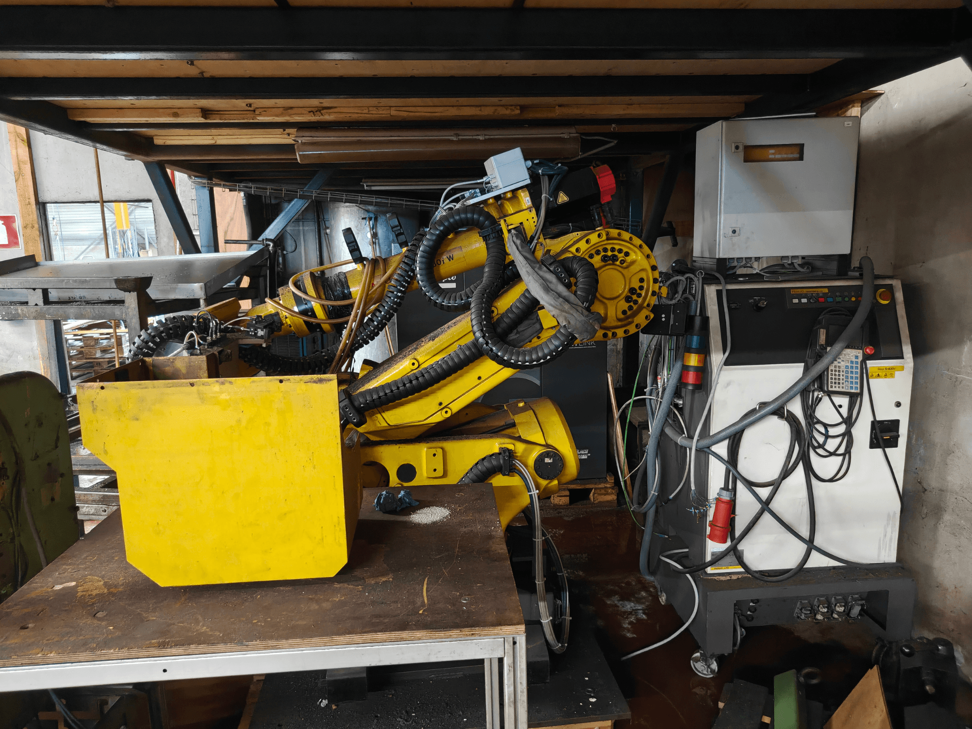 Vooraanzicht van een FANUC S-430i W-robotarm, geel, met bedieningspaneel, kabels en een geel onderdeel op een werkbank.