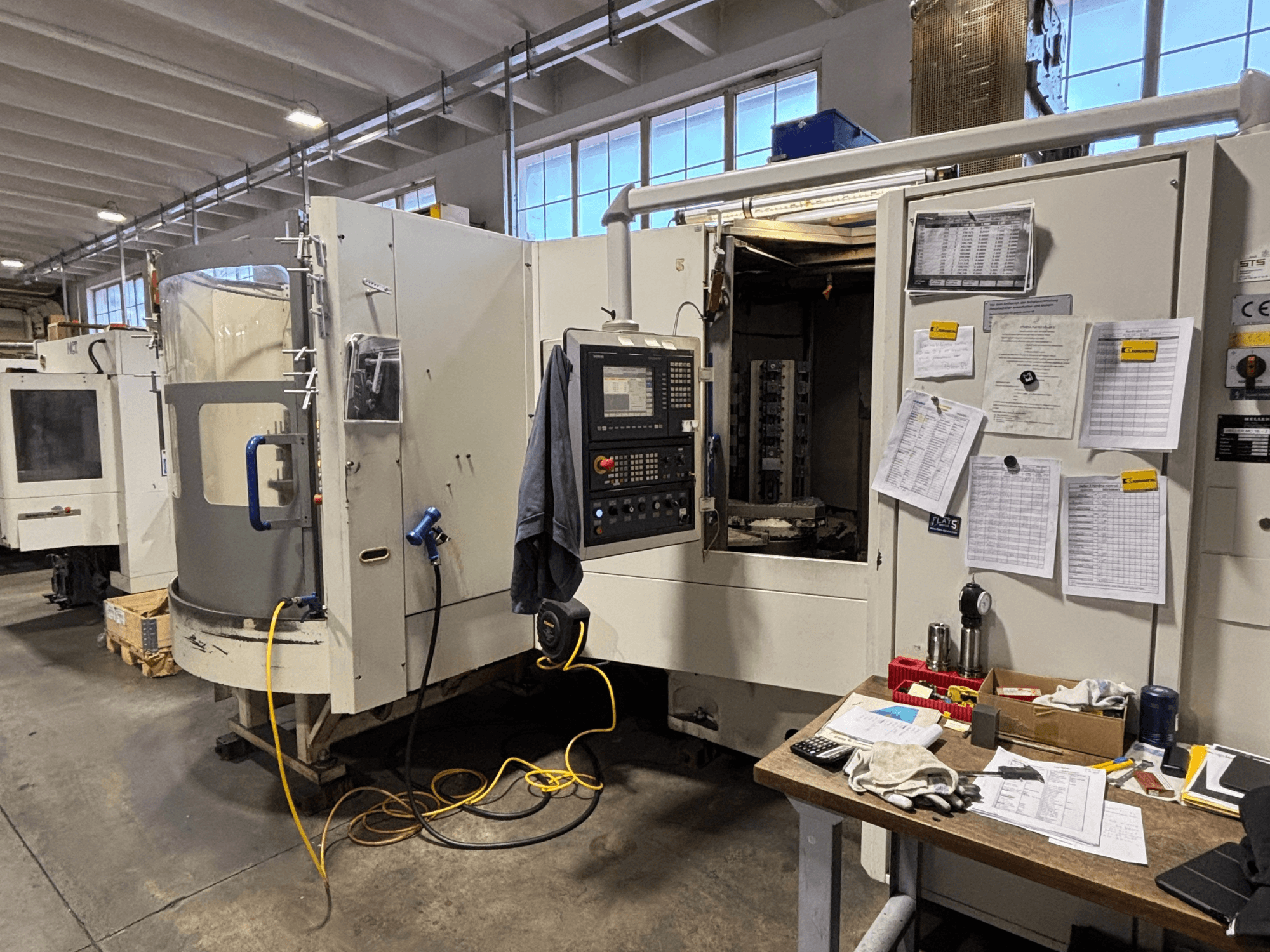 CNC machine in een werkplaats, vooraanzicht, met bedieningspaneel en documentatie aan de zijkant; gereedschappen en materialen op de tafel.