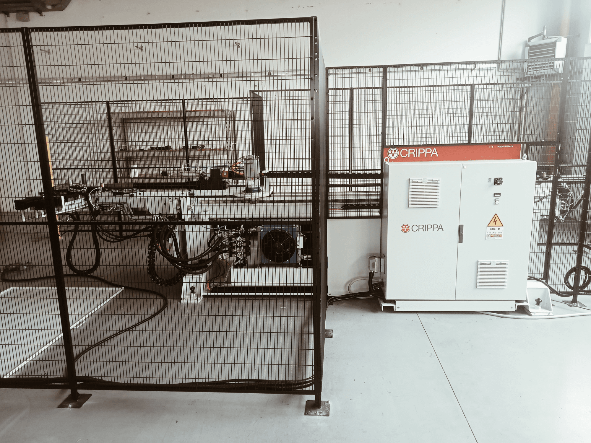 Witte CRIPPA machine achter een veiligheidskooi, met zichtbare slangen en onderdelen, zijaanzicht, industriële omgeving.