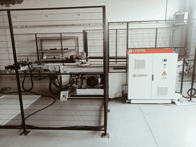 Witte CRIPPA machine achter een veiligheidskooi, met zichtbare slangen en onderdelen, zijaanzicht, industriële omgeving.