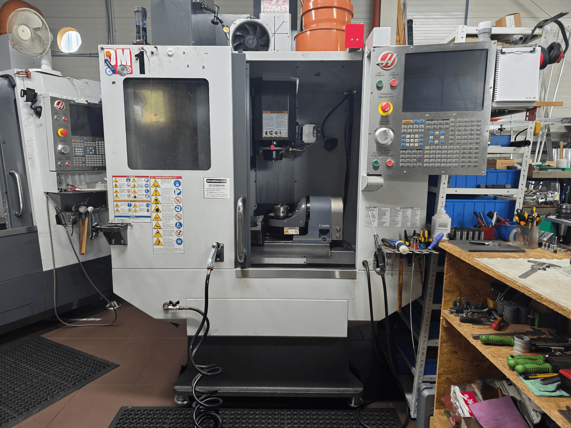 Haas CNC machine model VF-2, vooraanzicht, met bedieningselementen, spindelgebied, veiligheidslabels en gereedschappen op een nabijgelegen werkbank.