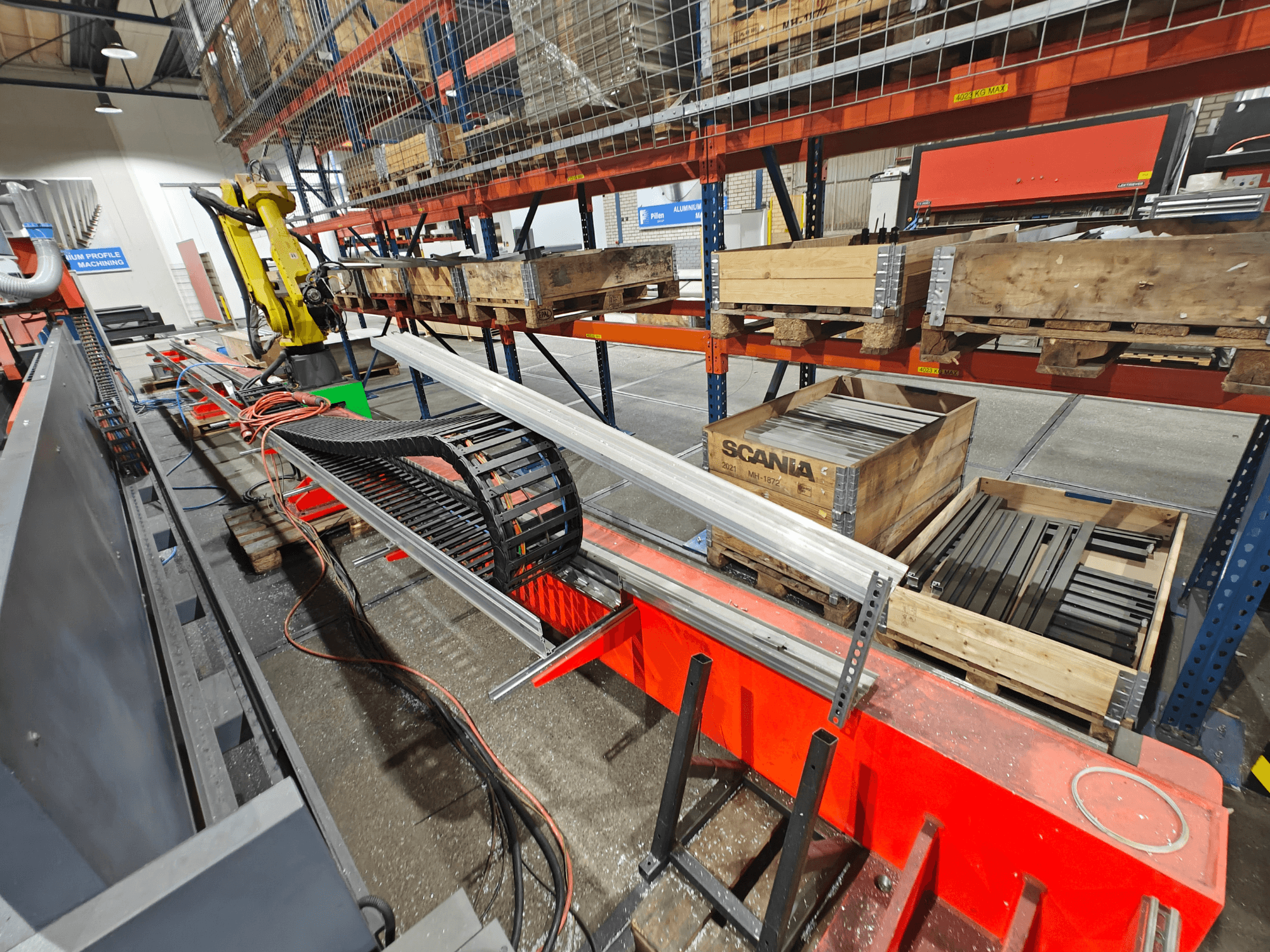 Robotarm op werkstation, oranje en zwarte kabels zichtbaar, met Scania-kratten en aluminium profielen in een fabrieksomgeving.