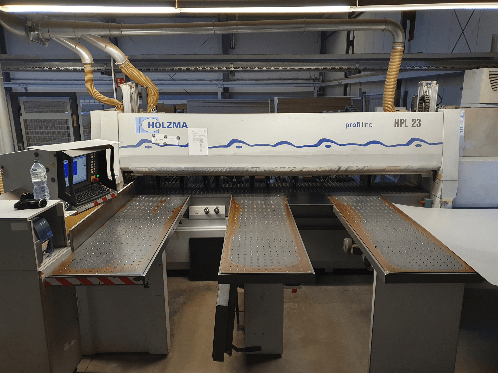 HOLZMA HPL 23 snijmachine in vooraanzicht, met drie geperforeerde tafels, computerterminal en waterfles in de buurt.