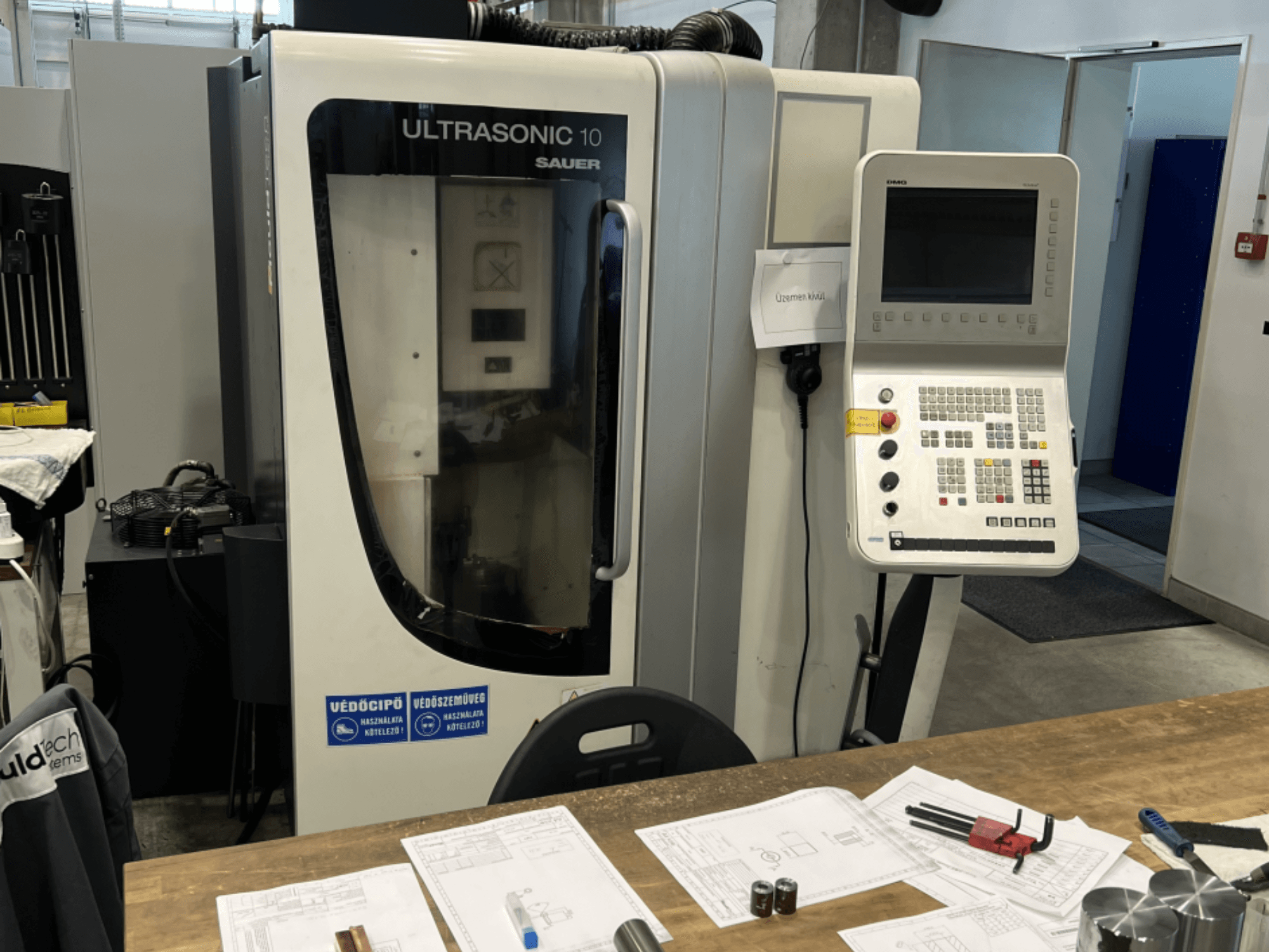 Vooraanzicht van DMG Ultrasonic 10 verticaal bewerkingscentrum met bedieningspaneel, met ontwerpdocumenten en gereedschappen op een tafel.