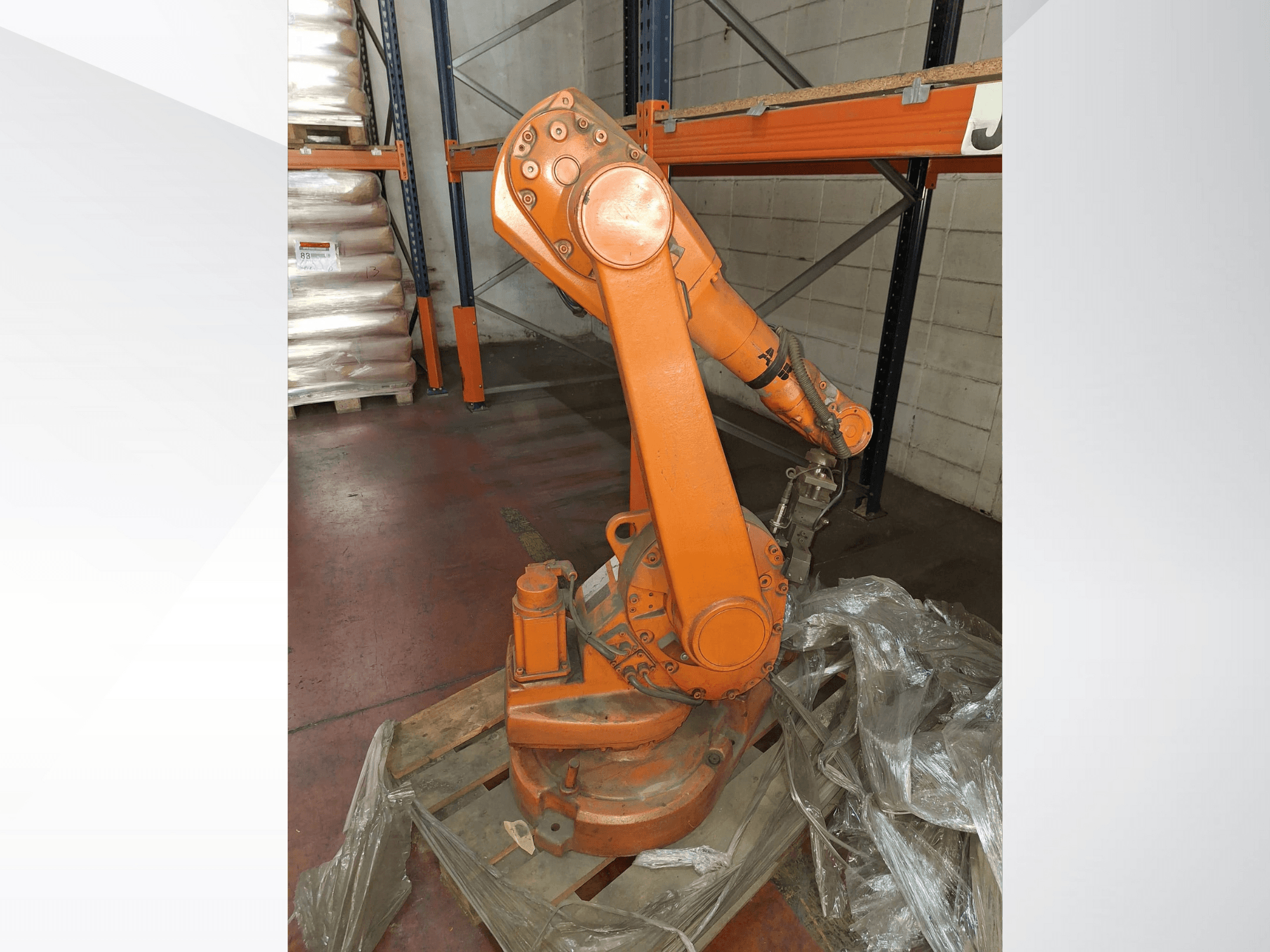Industriële robotarm in oranje, vooraanzicht op houten pallet, met bevestigde kabel en beschermend plastic omhulsel rond basis.