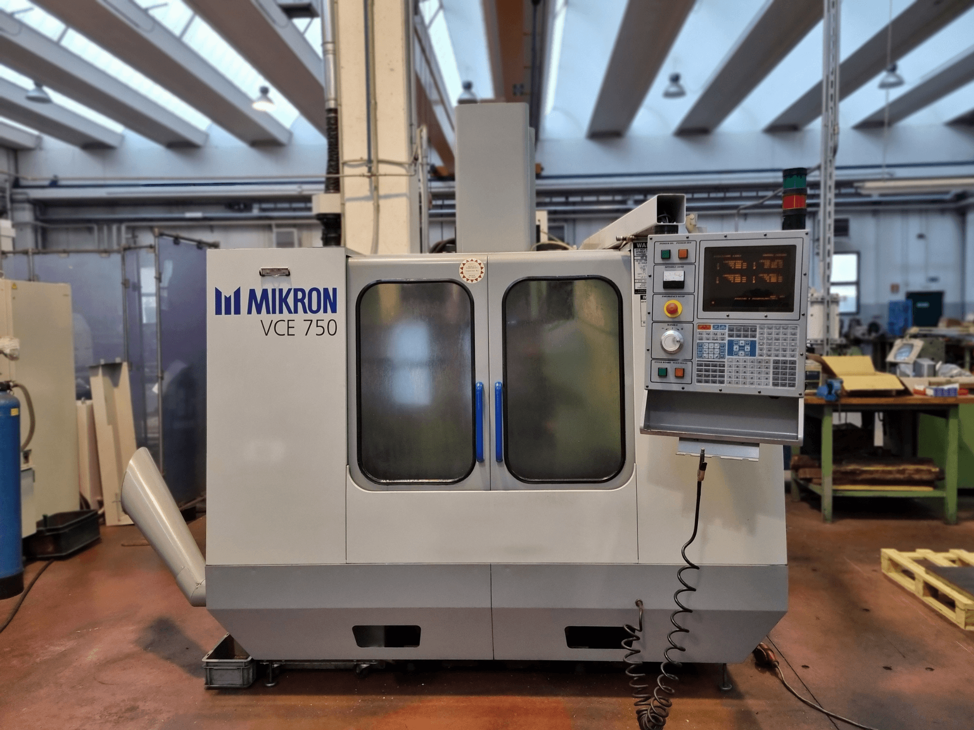 Witte Mikron VCE 750 CNC machine in een werkplaats, zijaanzicht, met een digitaal bedieningspaneel en grote glazen deuren.