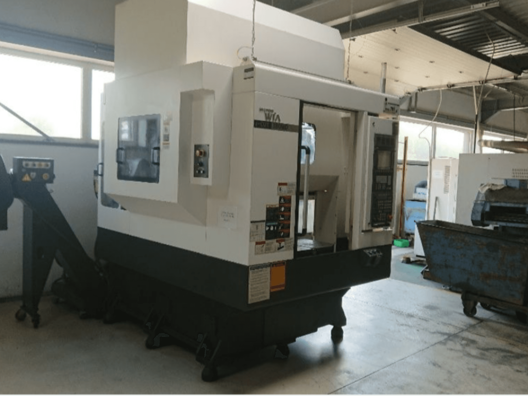 Haas CNC machine, zijaanzicht, met bedieningspaneel, waarschuwingslabels en een laadklep, opgesteld in een werkplaatsomgeving.
