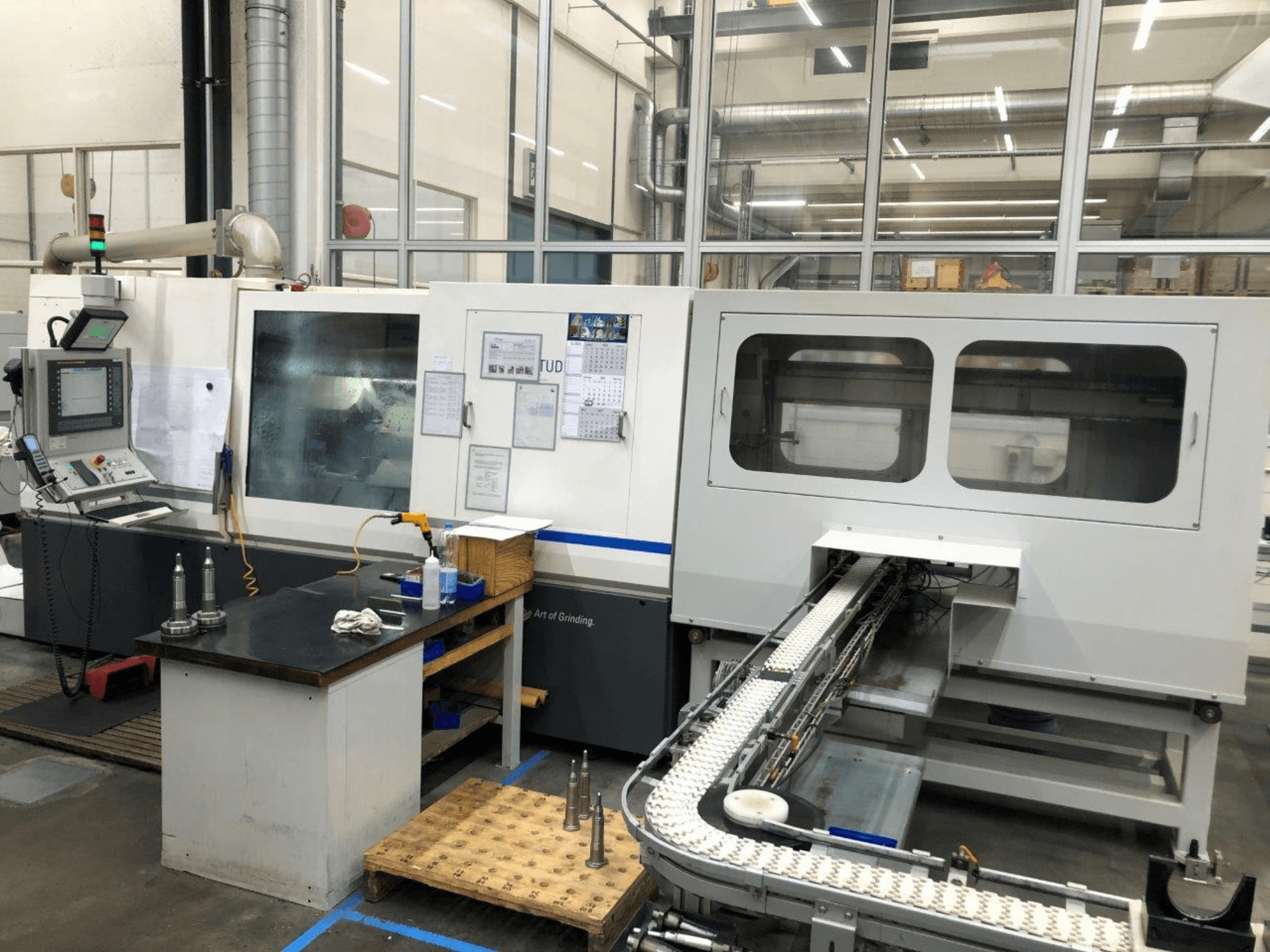CNC slijpmachine met bedieningspaneel, vooraanzicht; heeft een transportband en duidelijke kijkvensters, waarop precisiegereedschappen te zien zijn.