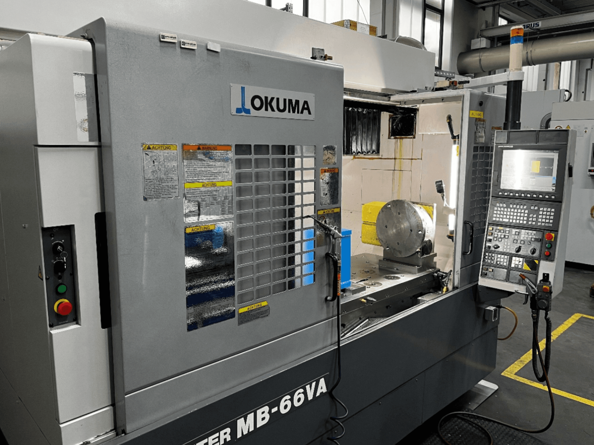 Okuma MB66-VA verticaal bewerkingscentrum vooraanzicht met bedieningspaneel, werkstuk en machine-interieur met veiligheidslabels.
