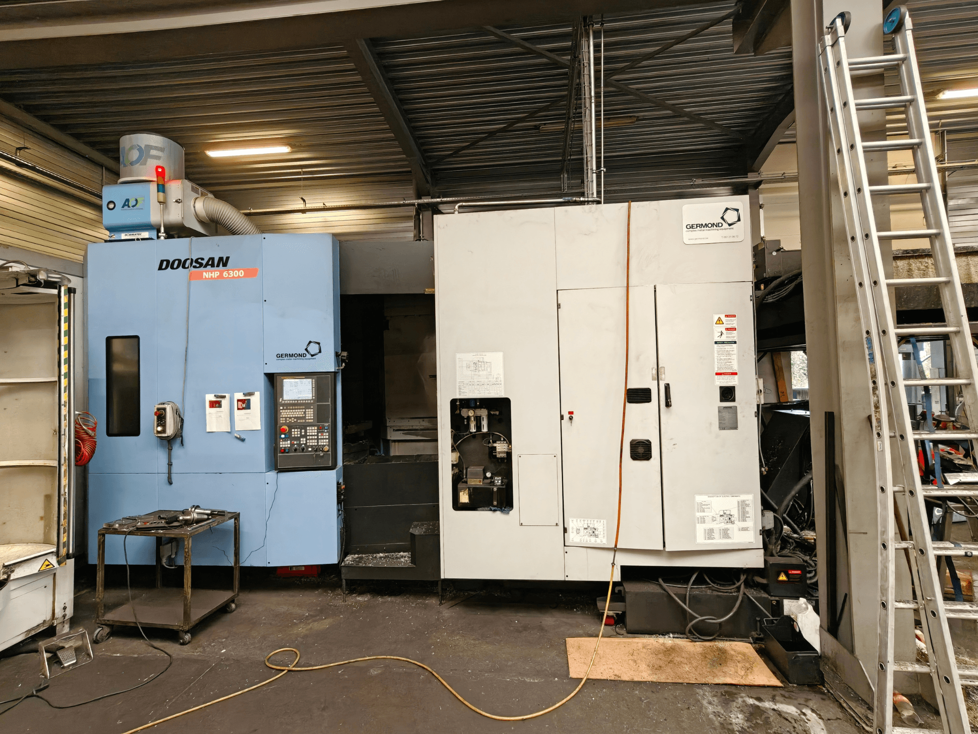 Het horizontale bewerkingscentrum DOOSAN NHP6300 in vooraanzicht, met het bedieningspaneel en de aangrenzende machines in een werkplaatsomgeving.