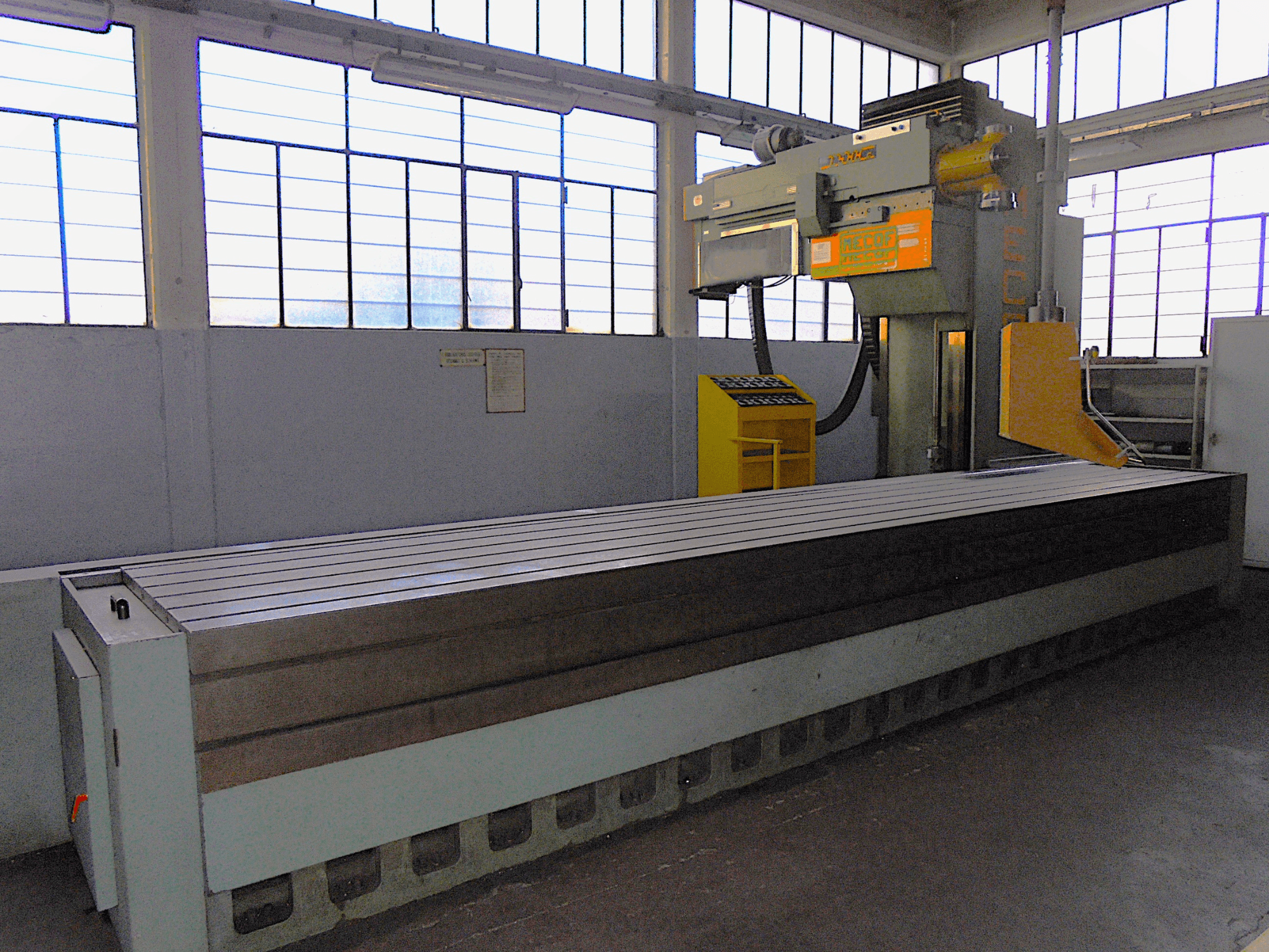 Merk onbekend, verticale freesmachine in een fabriek, zijaanzicht, met een grote tafel met gegroefd oppervlak en bedieningspaneel.