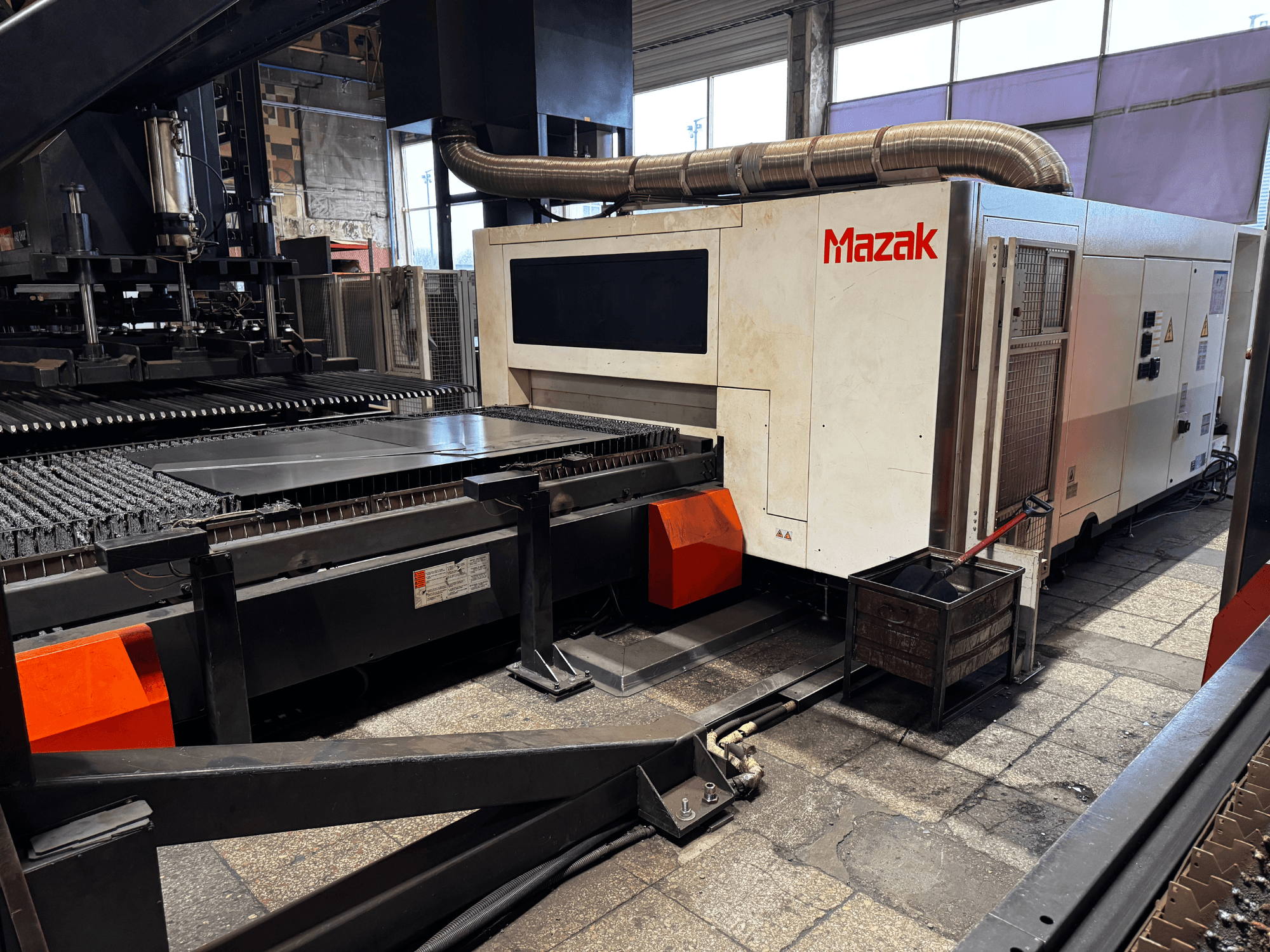 Mazak lasersnijmachine in een werkplaats, zijaanzicht met bedieningspaneel, transportband en een opvangbak.