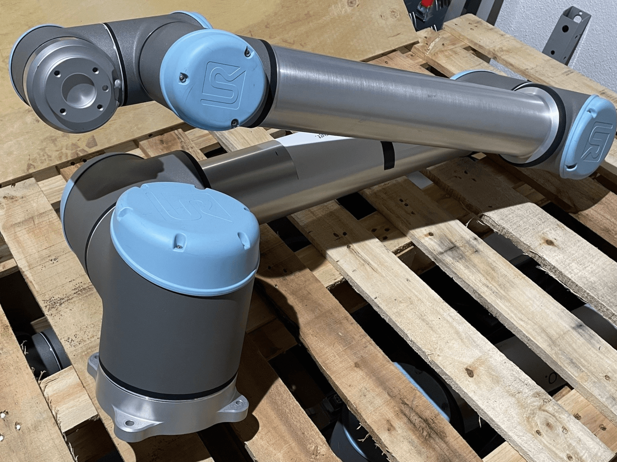 Zilveren robotarm met blauwe eindkappen, vooraanzicht, met gewrichten en mechanische details, rustend op een houten pallet.
