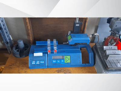 Blauwe Hommel T 1000 printer en T 1 010 tester met rode knoppen, gezien vanaf de voorkant op een werkbank met metalen blokken.