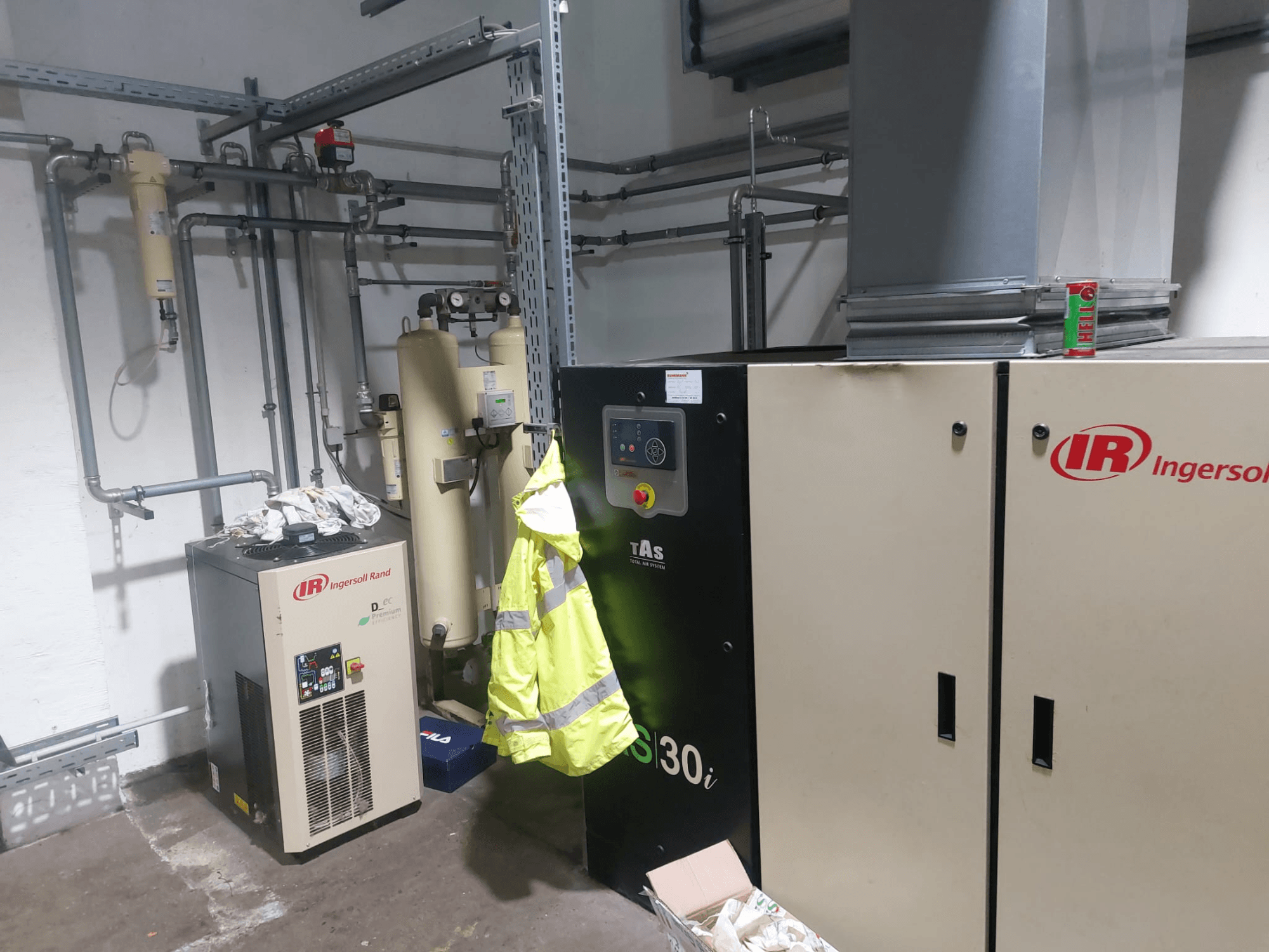Een Ingersoll Rand D480EC-A-bewerkingsmachine in een werkplaats, gezien vanaf de zijkant, met leidingen, bedieningselementen en een geel veiligheidsvest ernaast.