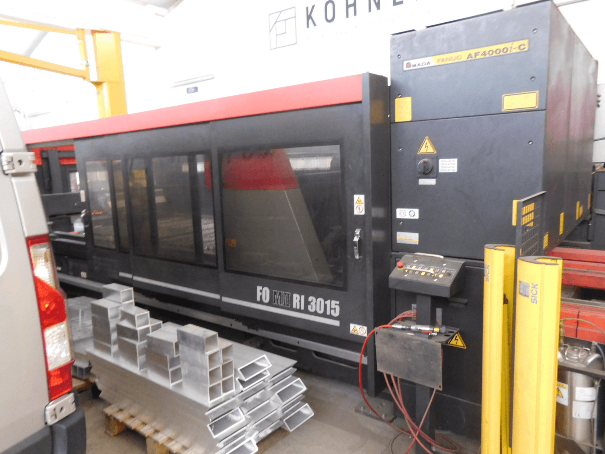 AMADA FO-3015 II RI CO2 lasersnijmachine in een werkplaats, zijaanzicht, met het bedieningspaneel en de gestapelde aluminium profielen.