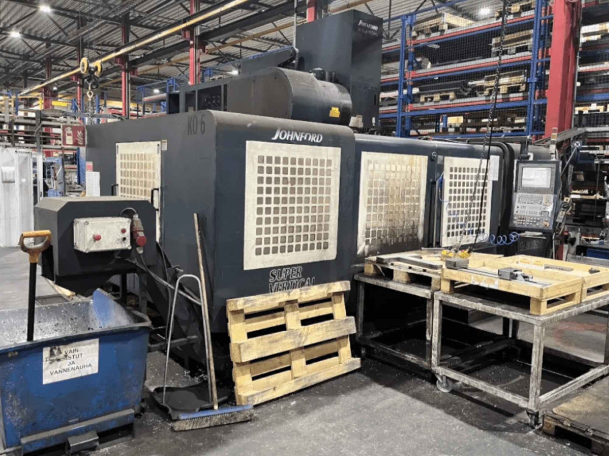 Johnford Super Vertical freesmachine in een fabriek, zijaanzicht met bedieningspaneel, pallets en reinigingsapparatuur in de buurt.