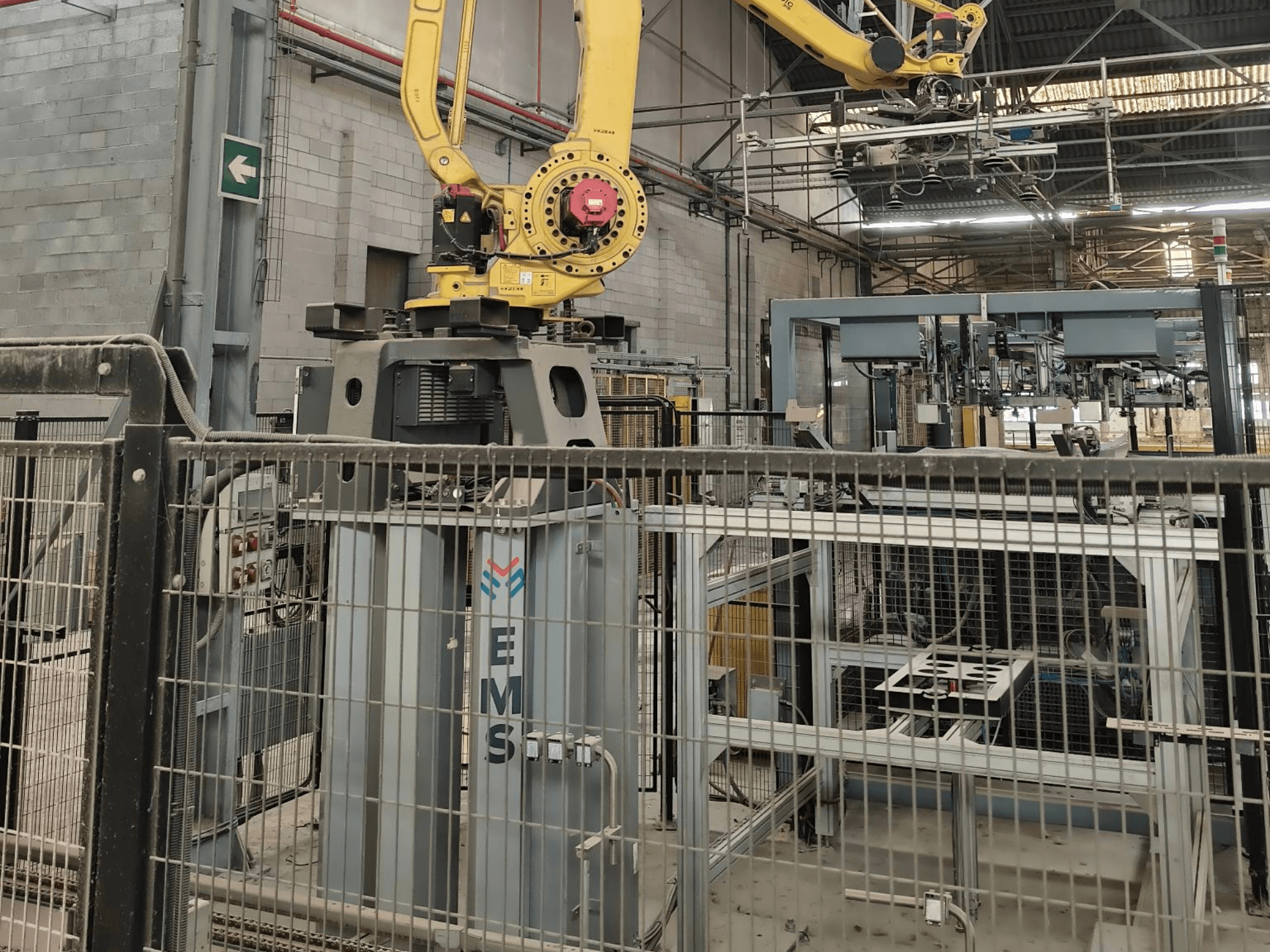 Vooraanzicht van een FANUC M-410iC/315-robotarm die boven een EMS-gereedschap is geplaatst, met industriële apparatuur en hekwerk op de achtergrond.