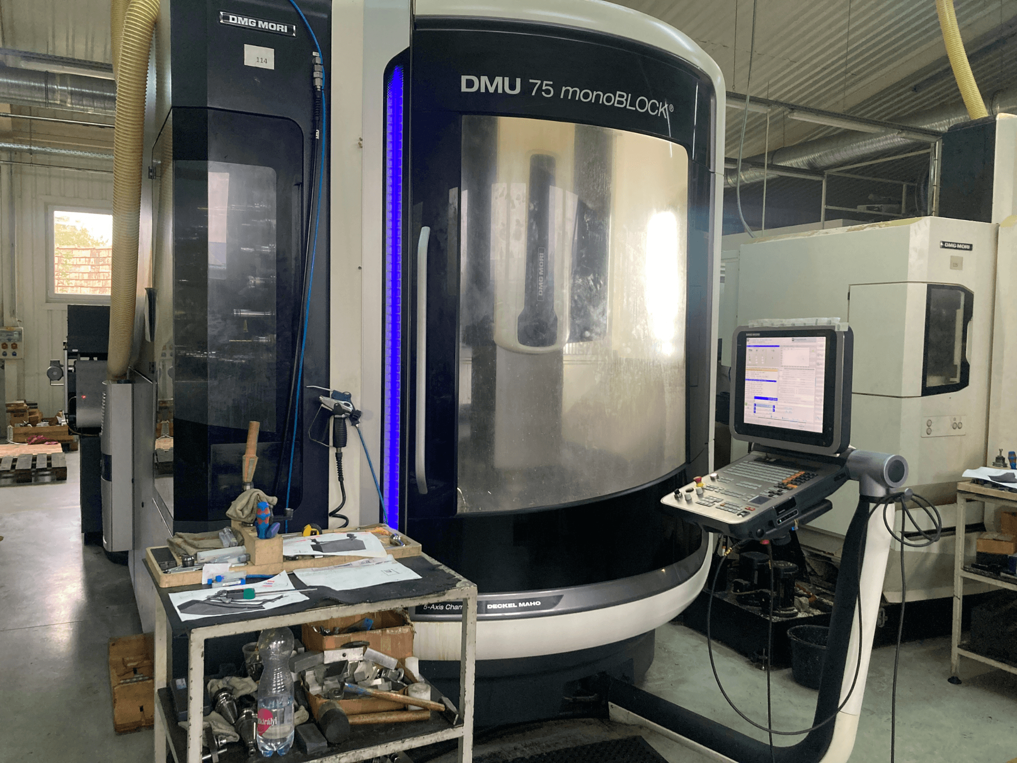 DMG MORI DMU 75 monoBLOCK® CNC machine, vooraanzicht, met bedieningspaneel, gereedschappen en een werkruimte met materialen.