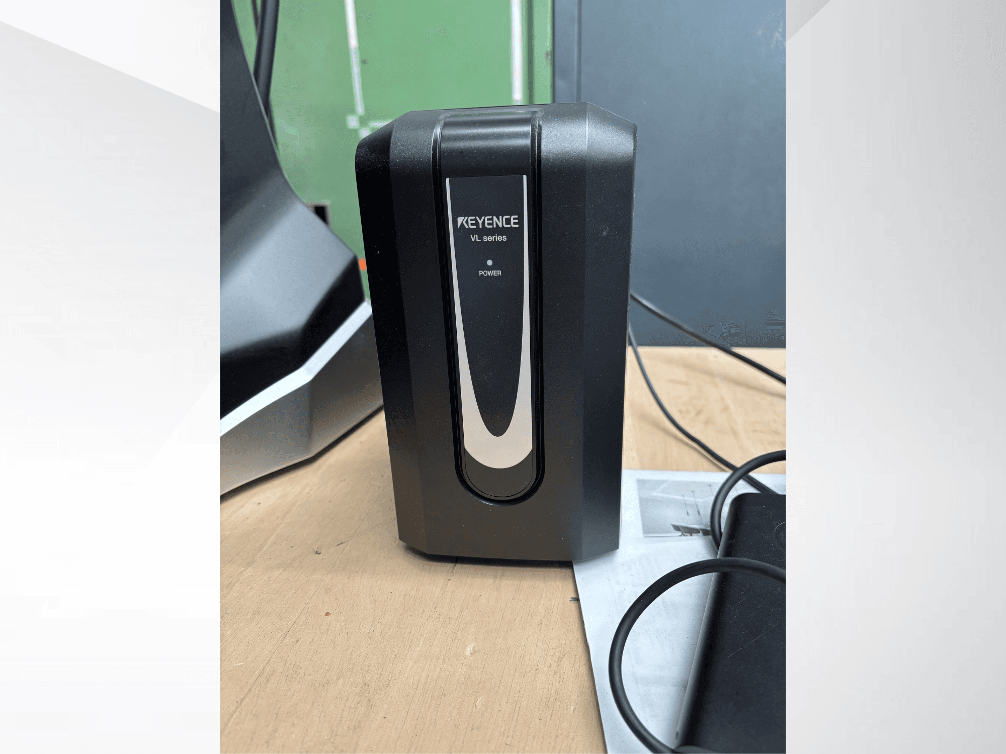 Zwarte KEYENCE VL-serie voedingseenheid, vooraanzicht, met stroomindicator en aangesloten kabels op een houten ondergrond.