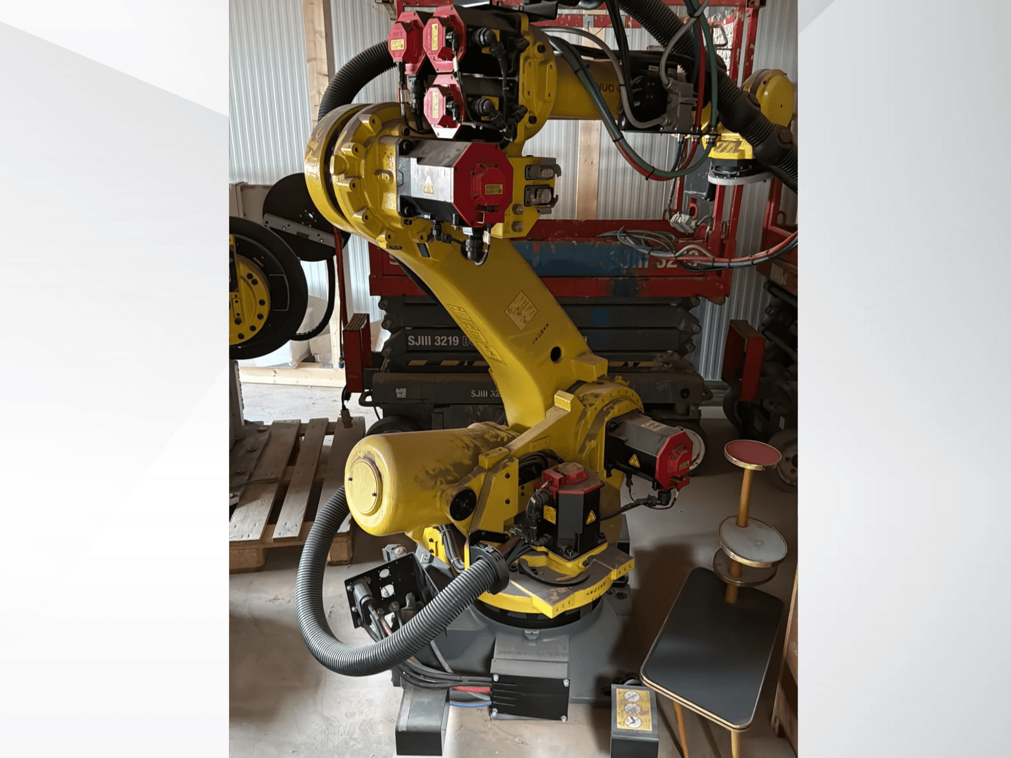 Gele industriële robotarm met rode controllers, vooraanzicht; voorzien van meerdere gewrichten en slangen, opgesteld op een bouwplaats.