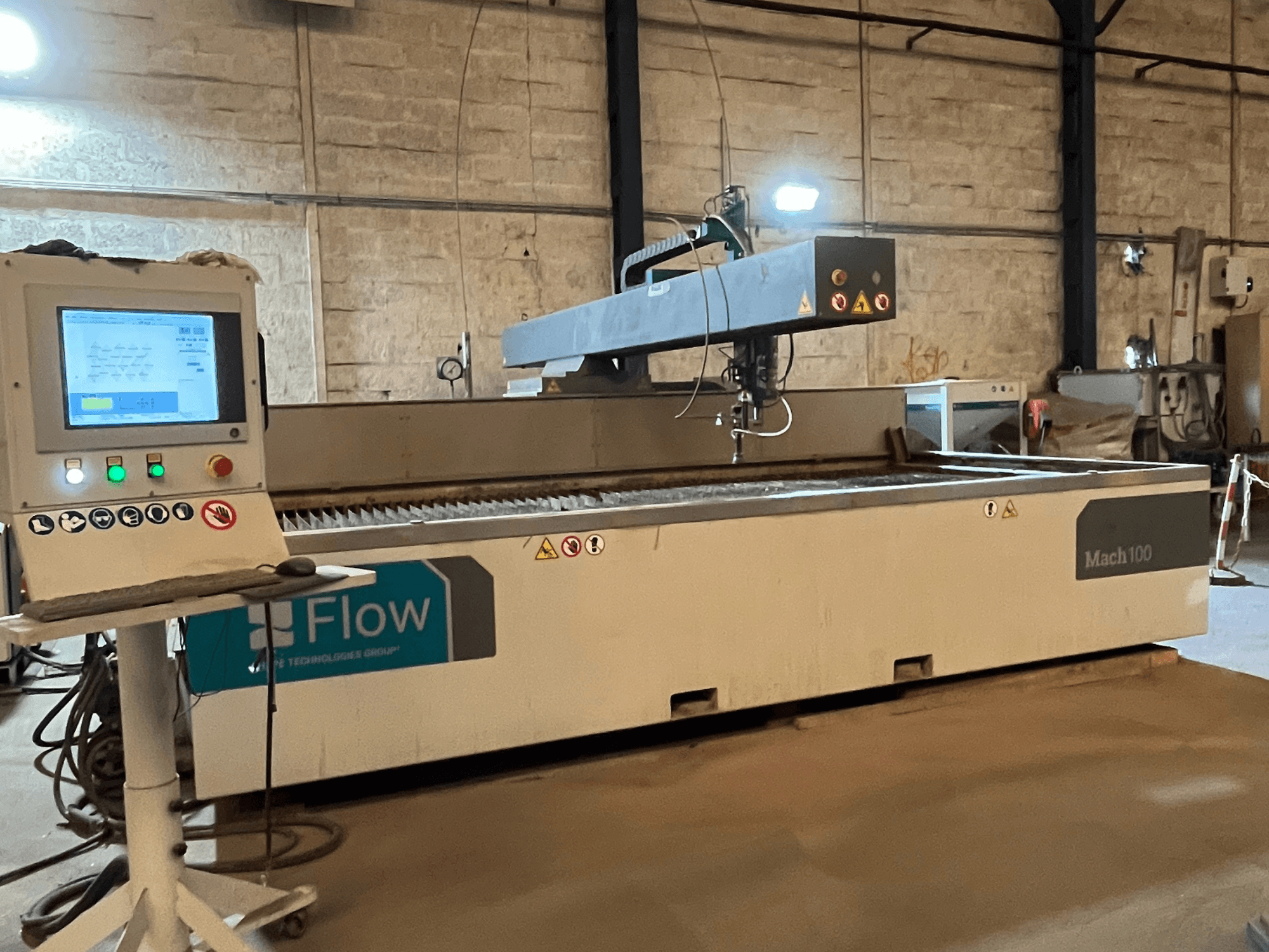 Flow Mach 100 waterstraalsnijder, zijaanzicht, met een digitaal bedieningspaneel en snijkop zichtbaar op een werktafel.
