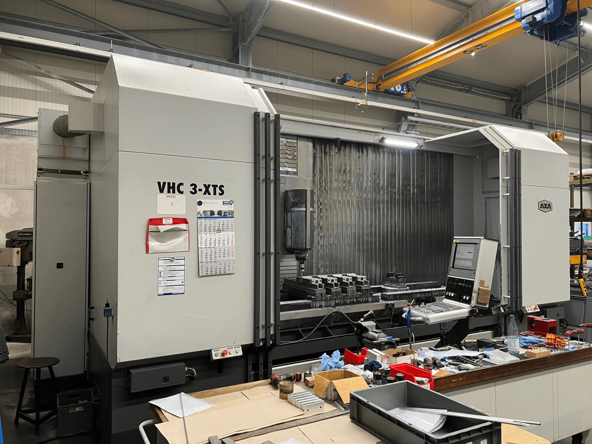 AXA VHC 3-XTS CNC machine in zijaanzicht, met bedieningspaneel en werkgebied vol gereedschap en onderdelen.