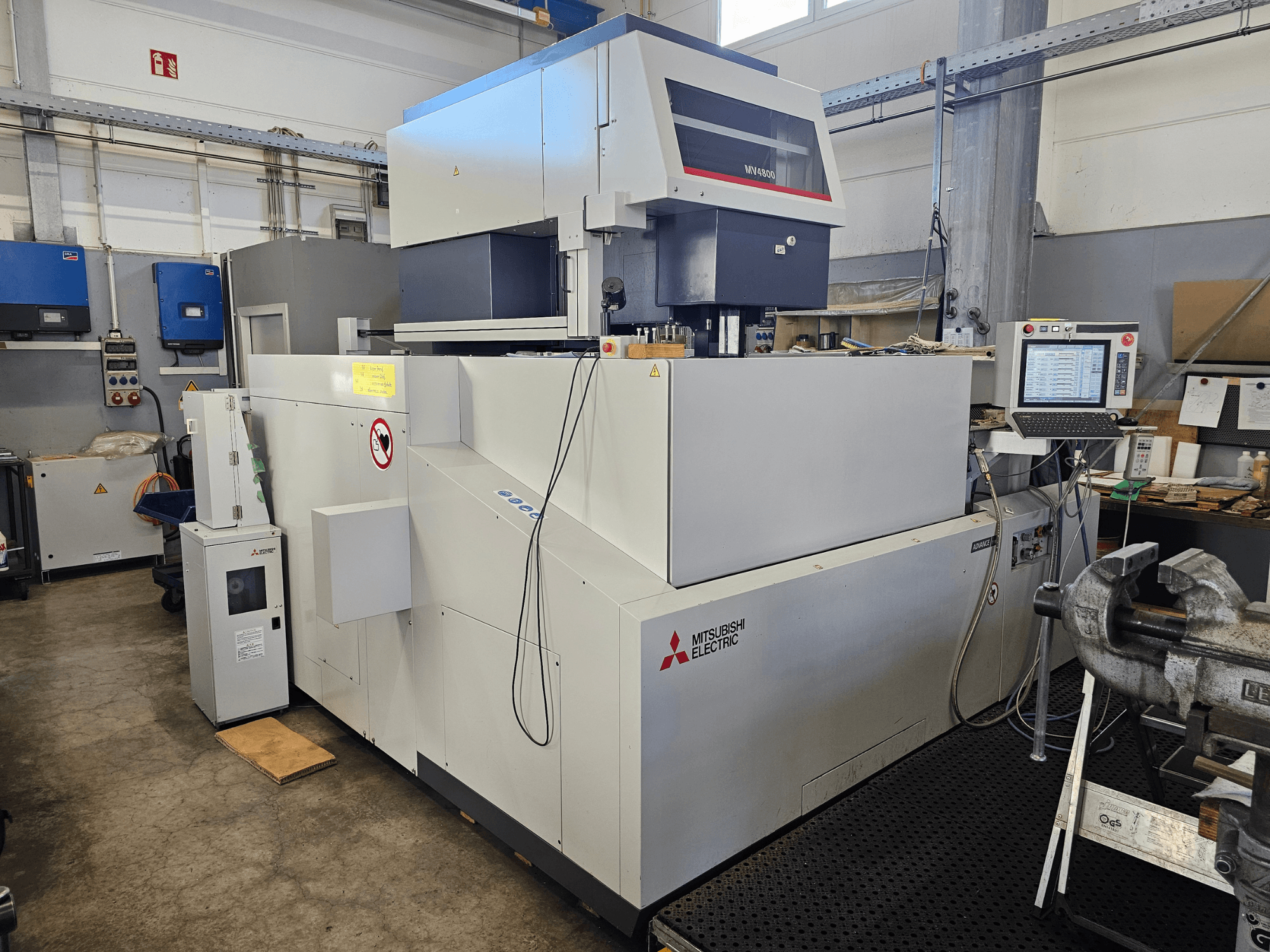 Mitsubishi MV4800 draadvonkmachine in een werkplaats, zijaanzicht met bedieningspaneel, veiligheidslabels en omringende apparatuur.