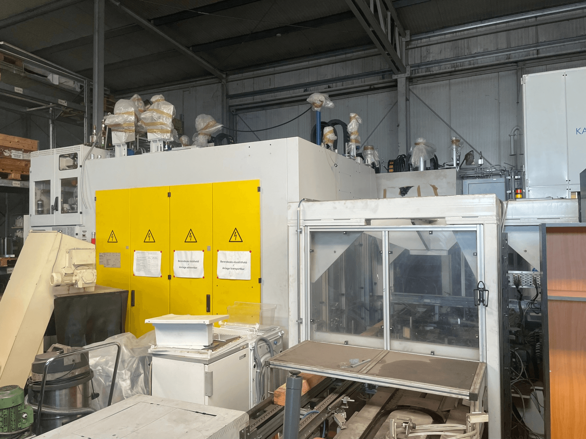 Industriële machines met geel bedieningspaneel, zijaanzicht, met waarschuwingslabels en plastic bekleding, in een magazijnomgeving.