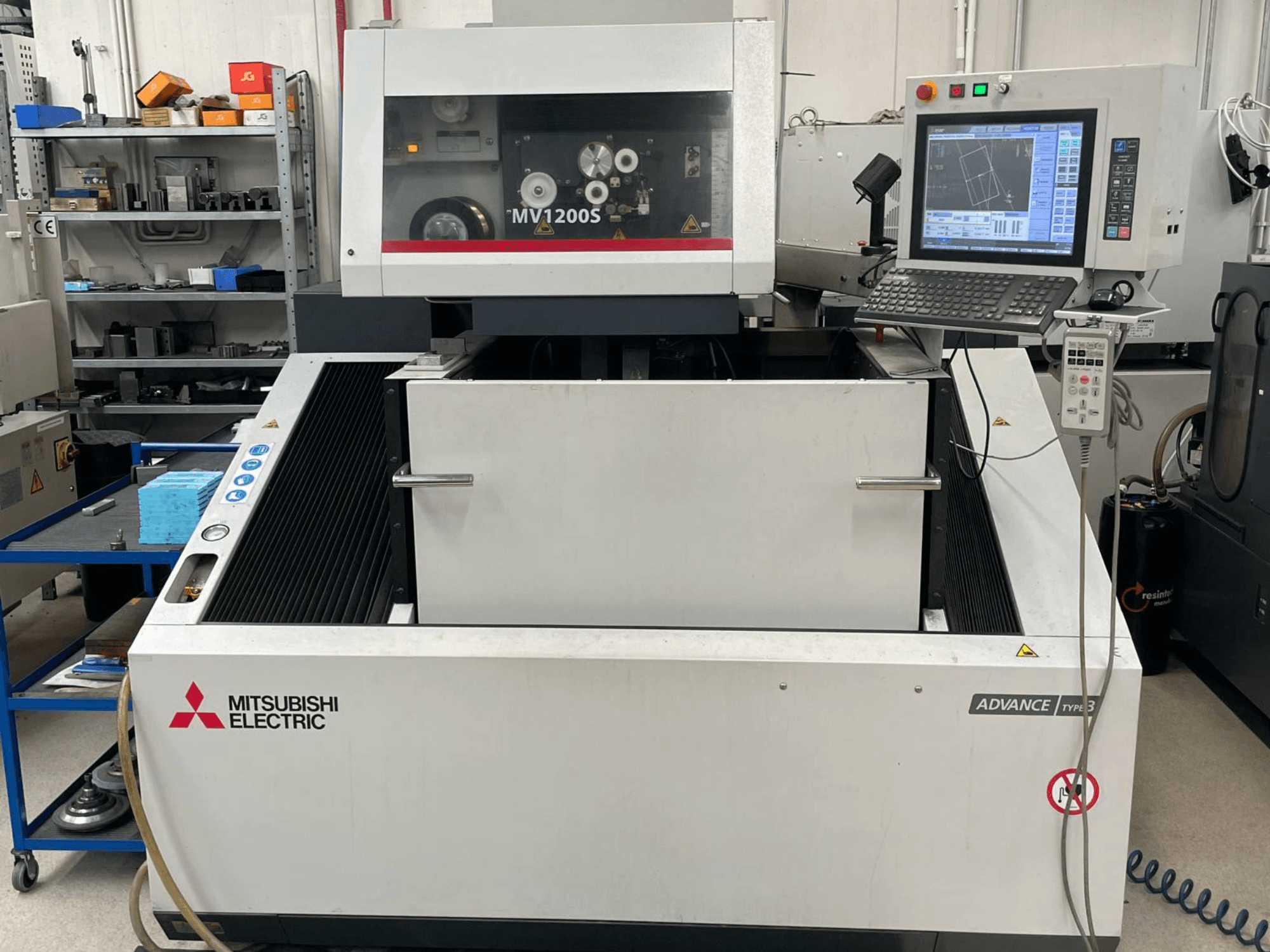 Mitsubishi MV1200S draadvonkmachine in vooraanzicht, met bedieningspaneel en werkgebied, waarbij de veiligheidswaarschuwingen zichtbaar zijn.