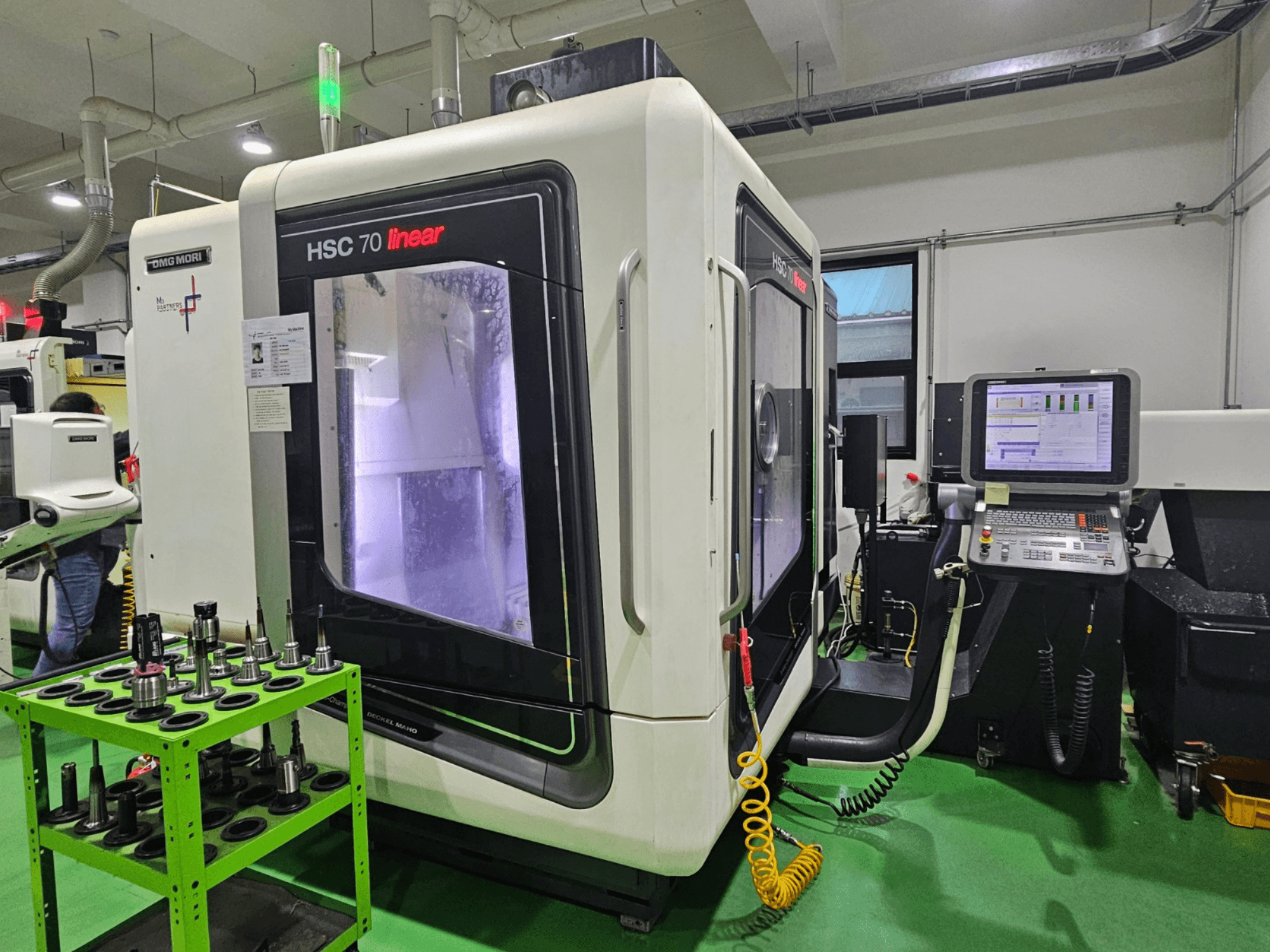 DMG MORI HSC 70 lineair universeel bewerkingscentrum, vooraanzicht, met gereedschapshouders op een groene tafel en een zichtbaar bedieningspaneel.