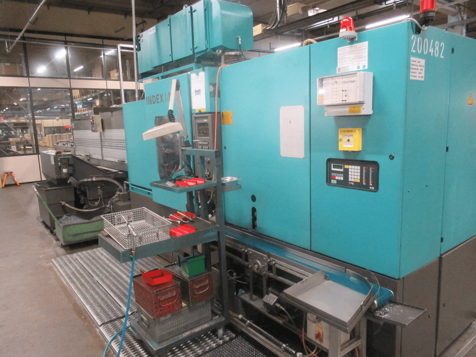 Turquoise INDEX machine in de fabriek, zijaanzicht; voorzien van bedieningspaneel, gereedschapopslag en transportband. Zichtbare bakken, slangen en alarmen.