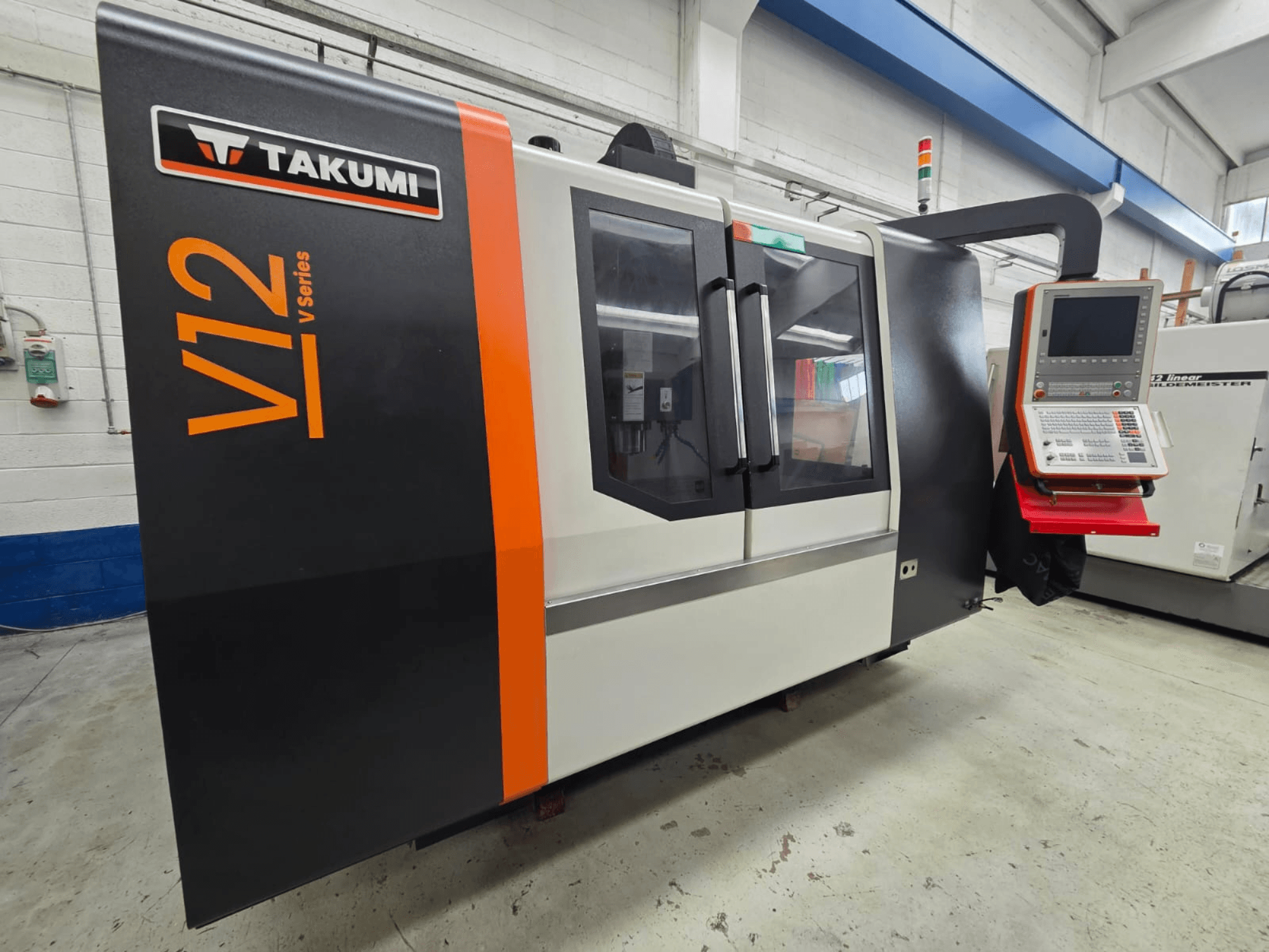 Takumi V12 V serie CNC freesmachine, vooraanzicht, heeft een bedieningspaneel en grote glazen vensters.