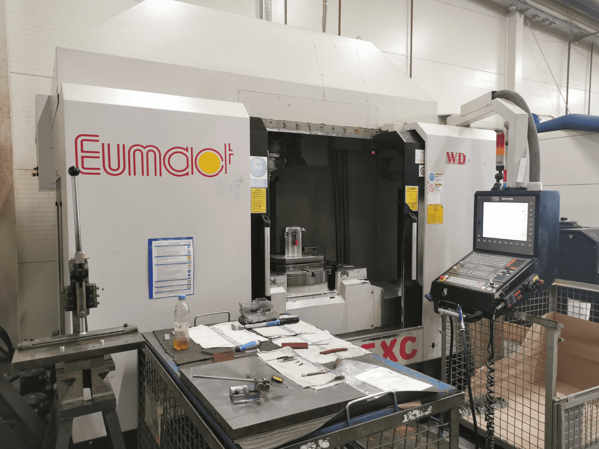 Vooraanzicht van een Eumdaot CNC machine met bedieningspaneel, gereedschappen en een werkoppervlak vol componenten en materialen.