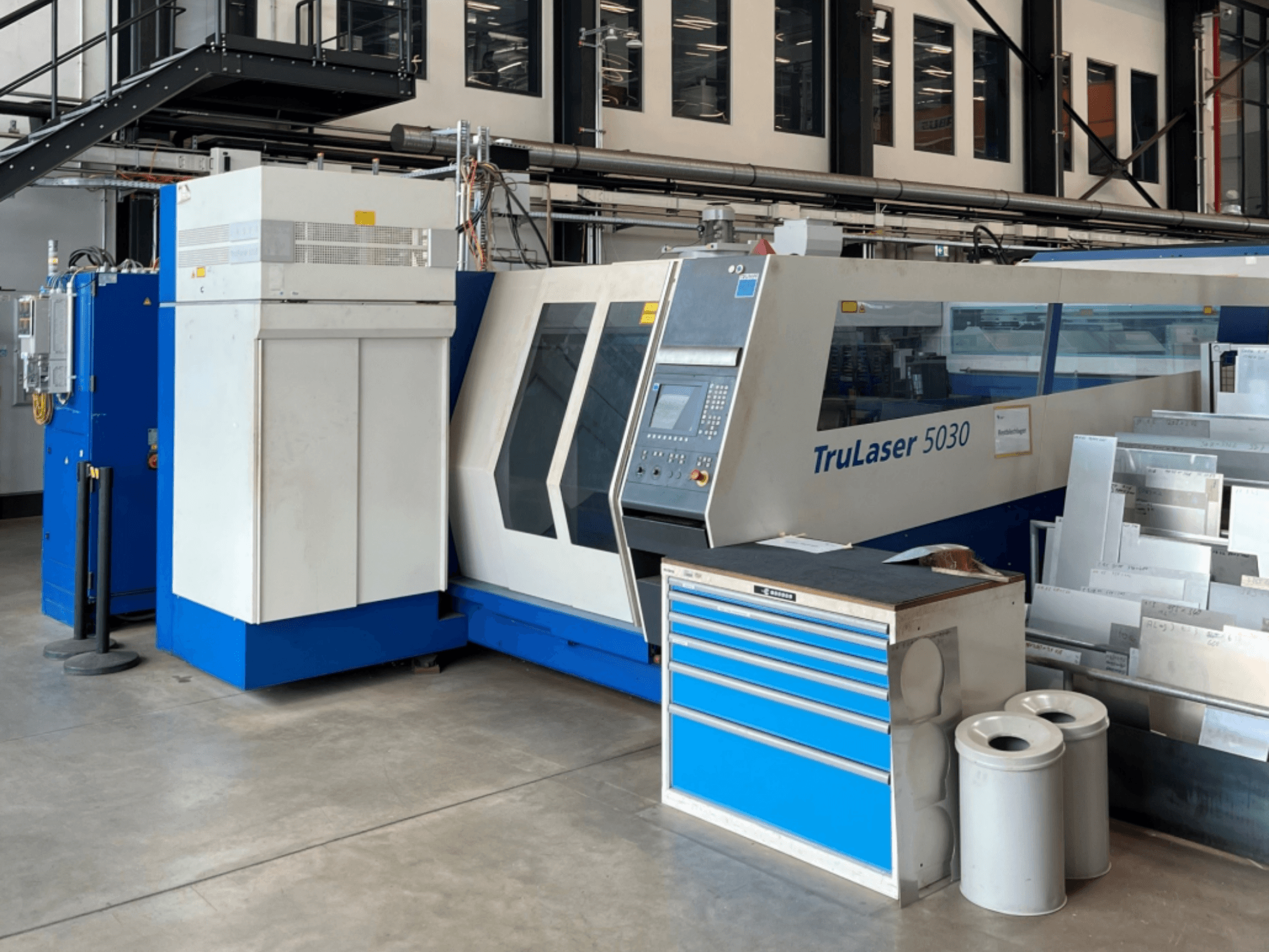 TRUMPF TruLaser 5030 CO₂-lasersnijmachine in vooraanzicht, met bedieningspaneel en een werkbank met metalen platen ernaast.