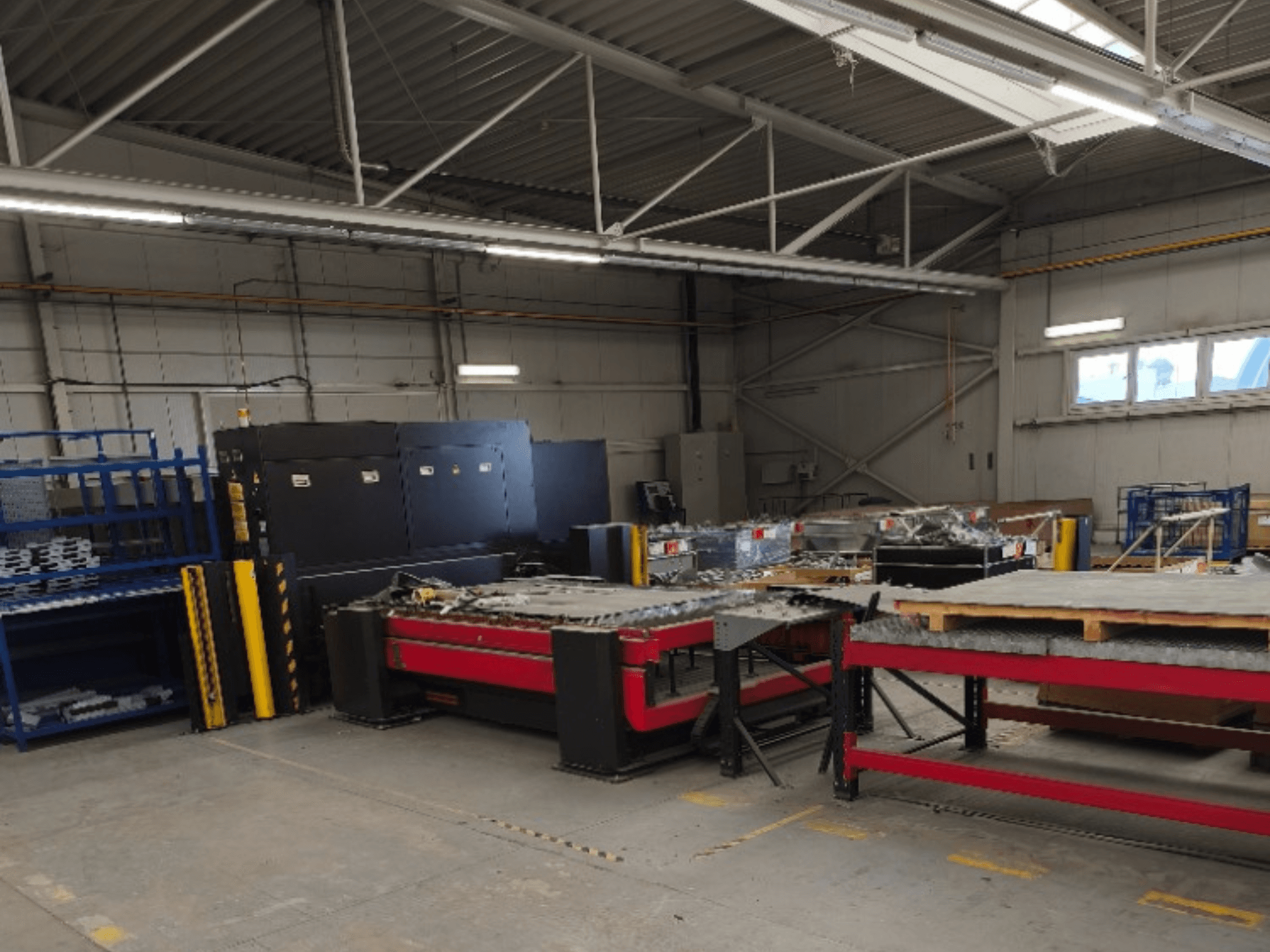 Een AMADA LCG 3015 AJ-vezellaser-snijmachine in een werkplaats, gezien vanaf de voorkant, met opbergrekken en materialen op de achtergrond.