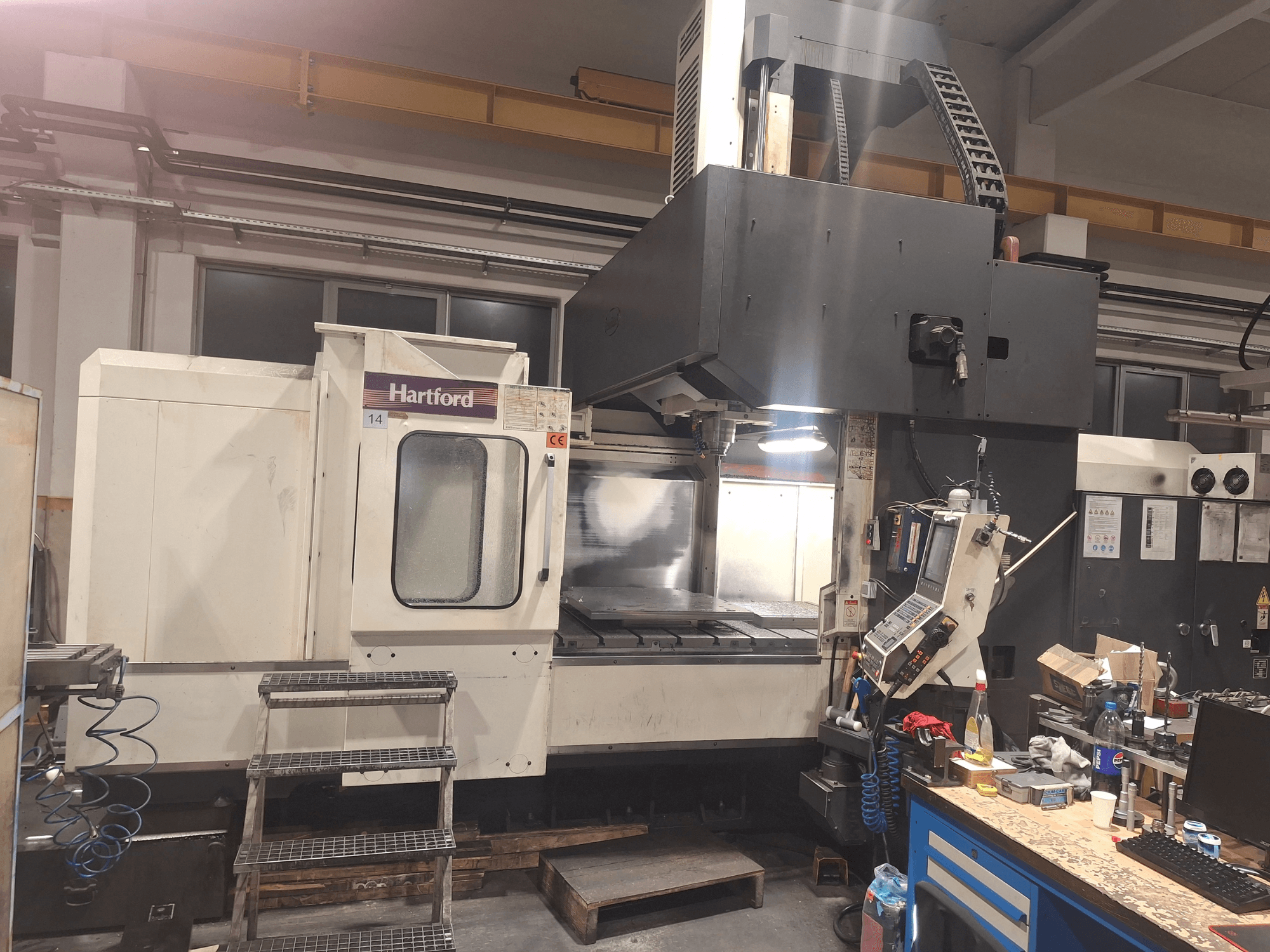Hartford CNC machine, vooraanzicht, met technische bedieningselementen, toegangstreden en een werktafel, in een werkplaats.