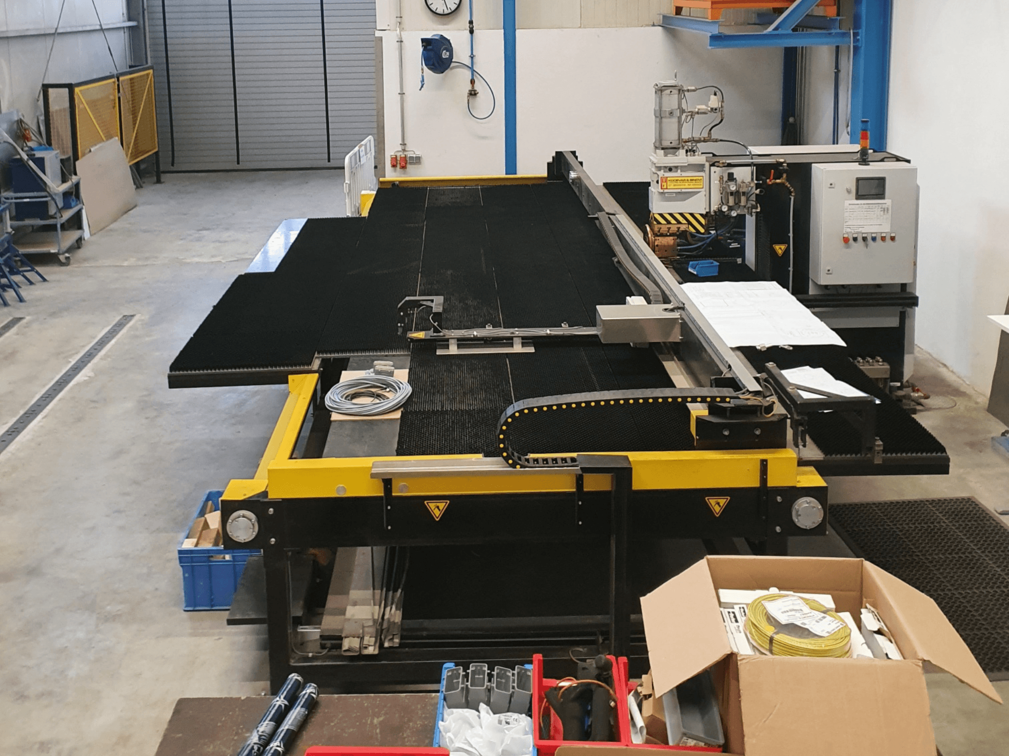 Industriële snijmachine met gele en zwarte kenmerken, vooraanzicht, waarop de bedieningselementen en accessoires in een fabrieksomgeving te zien zijn.