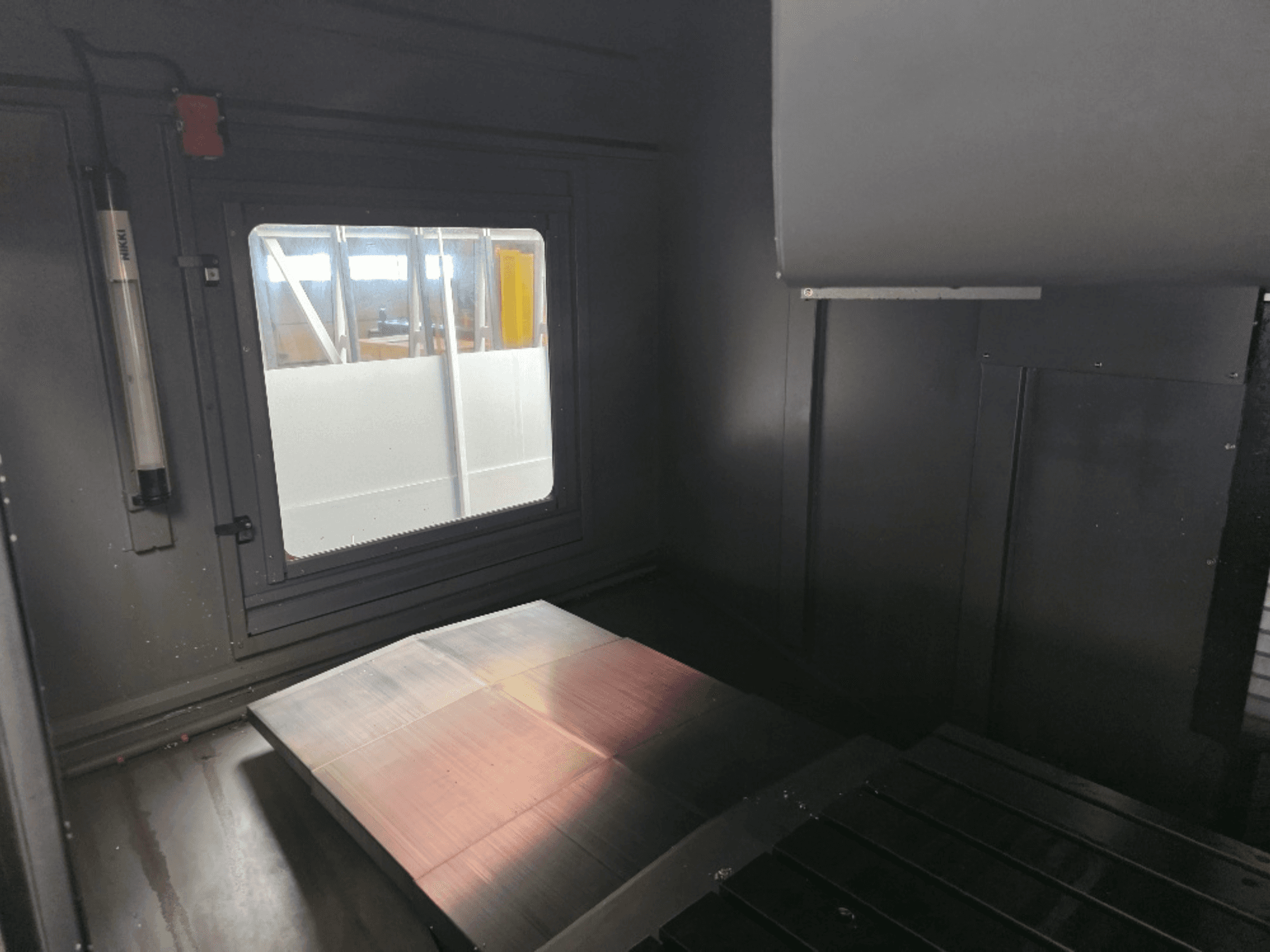 Binnenzicht van een machinebehuizing met een gepolijste metalen plaat, een zijraam en minimale verlichting. Geen merk zichtbaar.