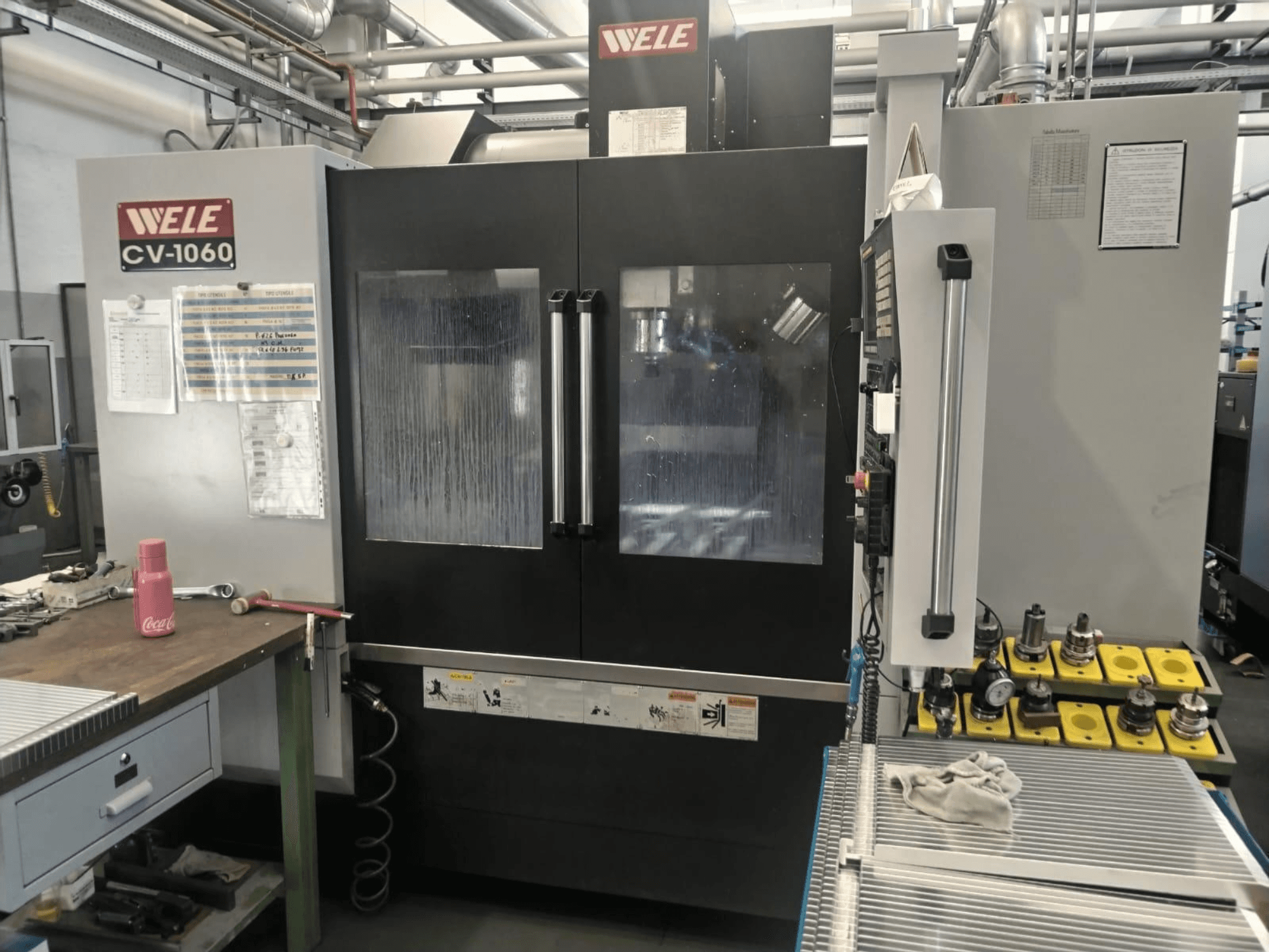 Wele CV-1060 verticaal bewerkingscentrum, vooraanzicht, met bedieningspaneel en zichtbaar werkgebied; inclusief hulpgereedschap en veiligheidslabels.