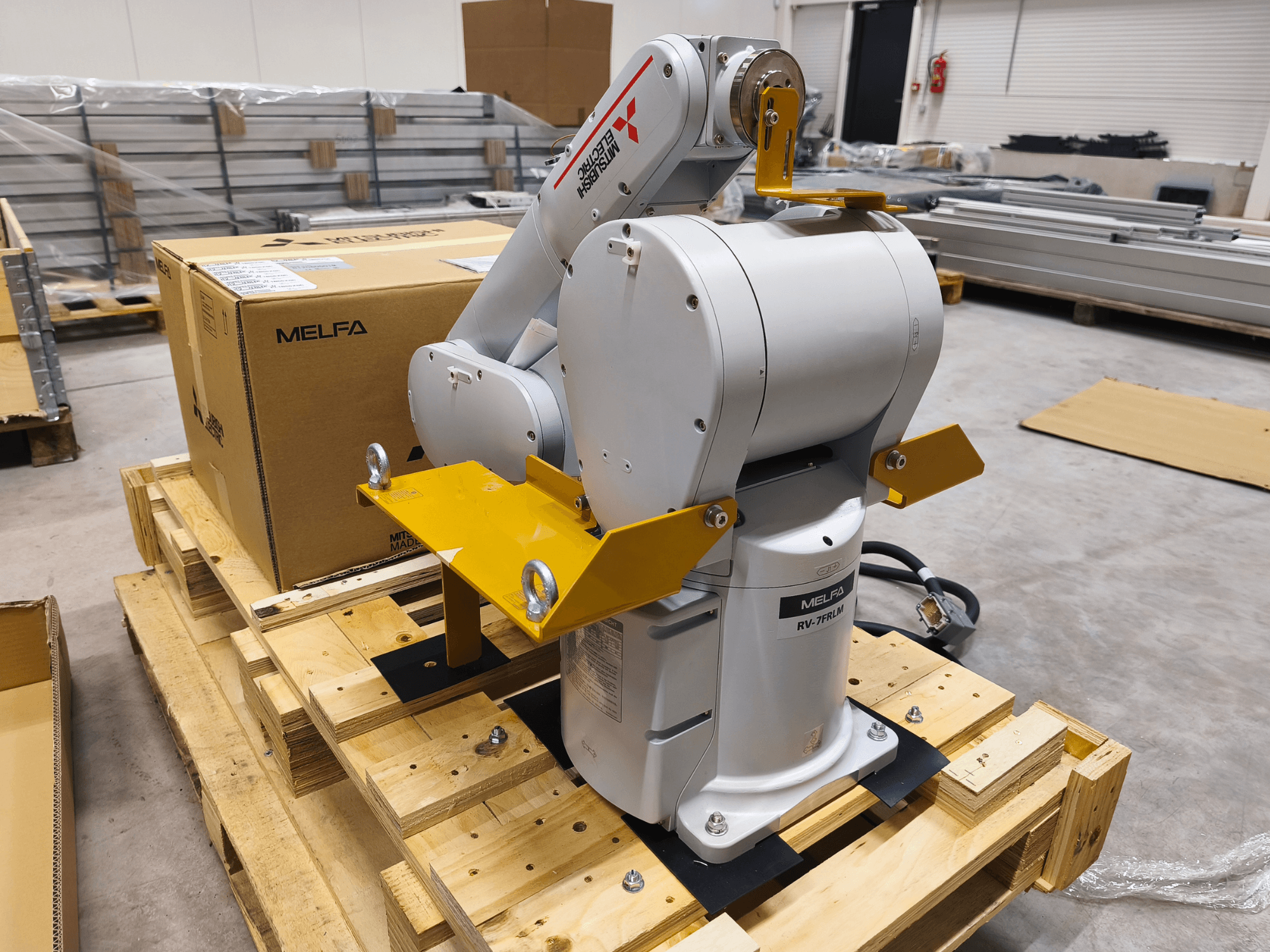 MELFA RV-7FLM industriële robotarm op houten pallet, vooraanzicht, met gele steunbeugels en grijze verpakkingsdoos.