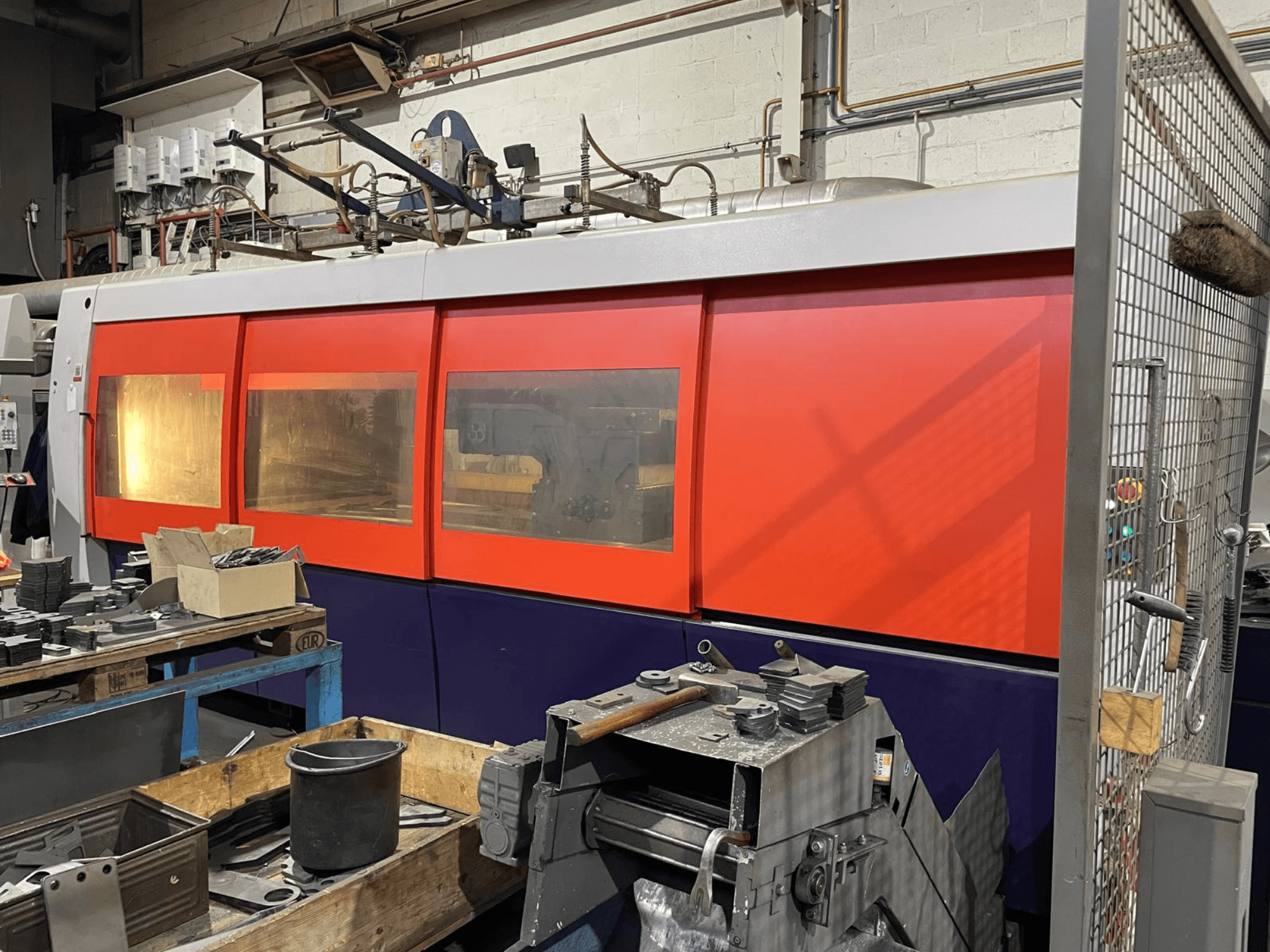 Close-up van een oranje en witte industriële lasersnijmachine, zijaanzicht, met bedieningspaneel en werkplaatsgereedschap op de voorgrond.