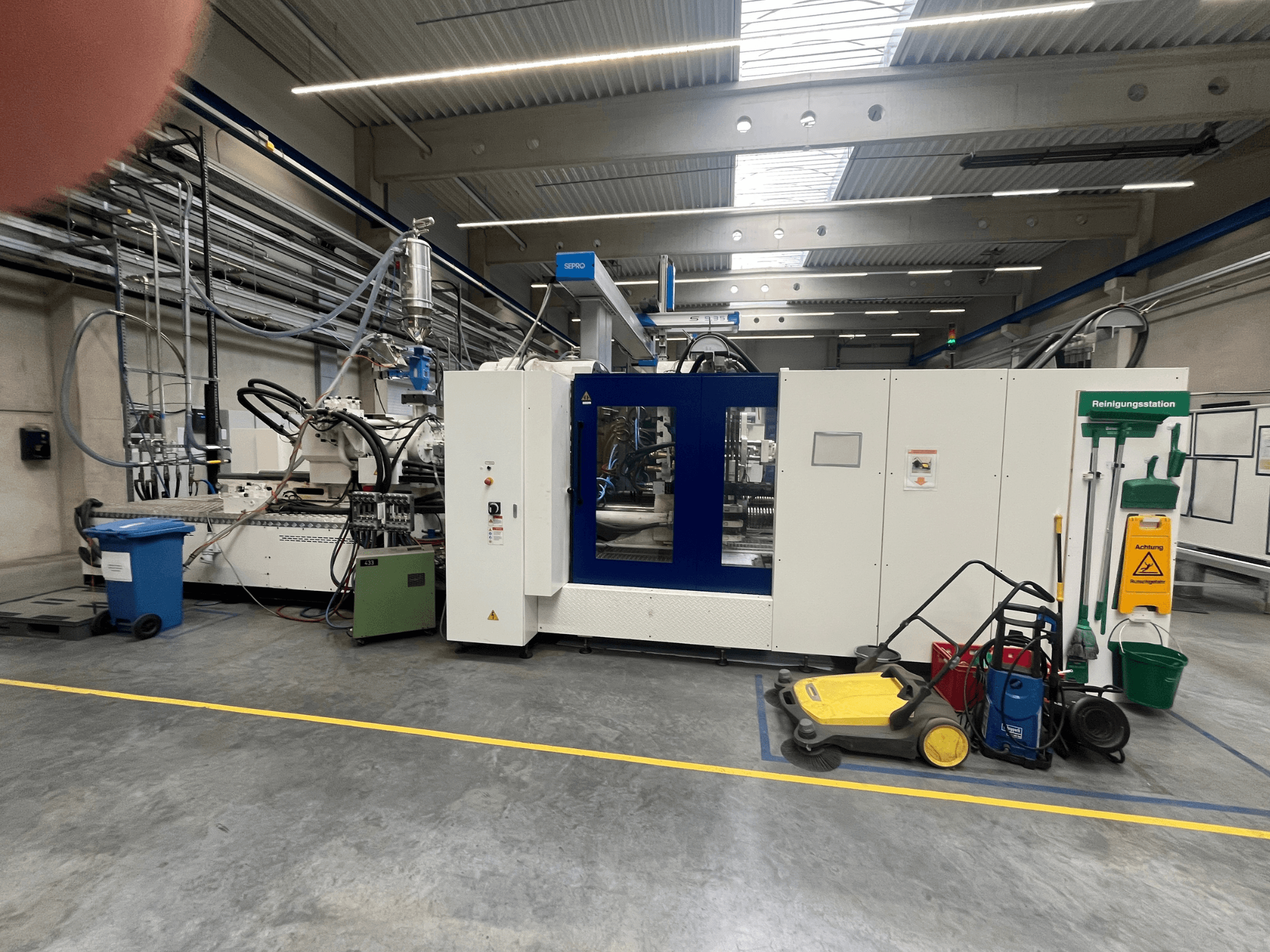 Industriële machine in een fabriek, vooraanzicht, met blauwe en witte panelen, slangen en reinigingsapparatuur in de buurt.