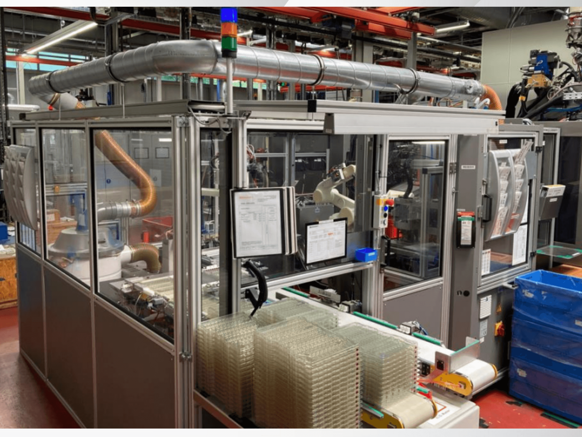 Vooraanzicht van een geautomatiseerde machine met metalen buizen, bedieningspaneel en gestapelde doorzichtige plastic bakken op een fabrieksvloer.