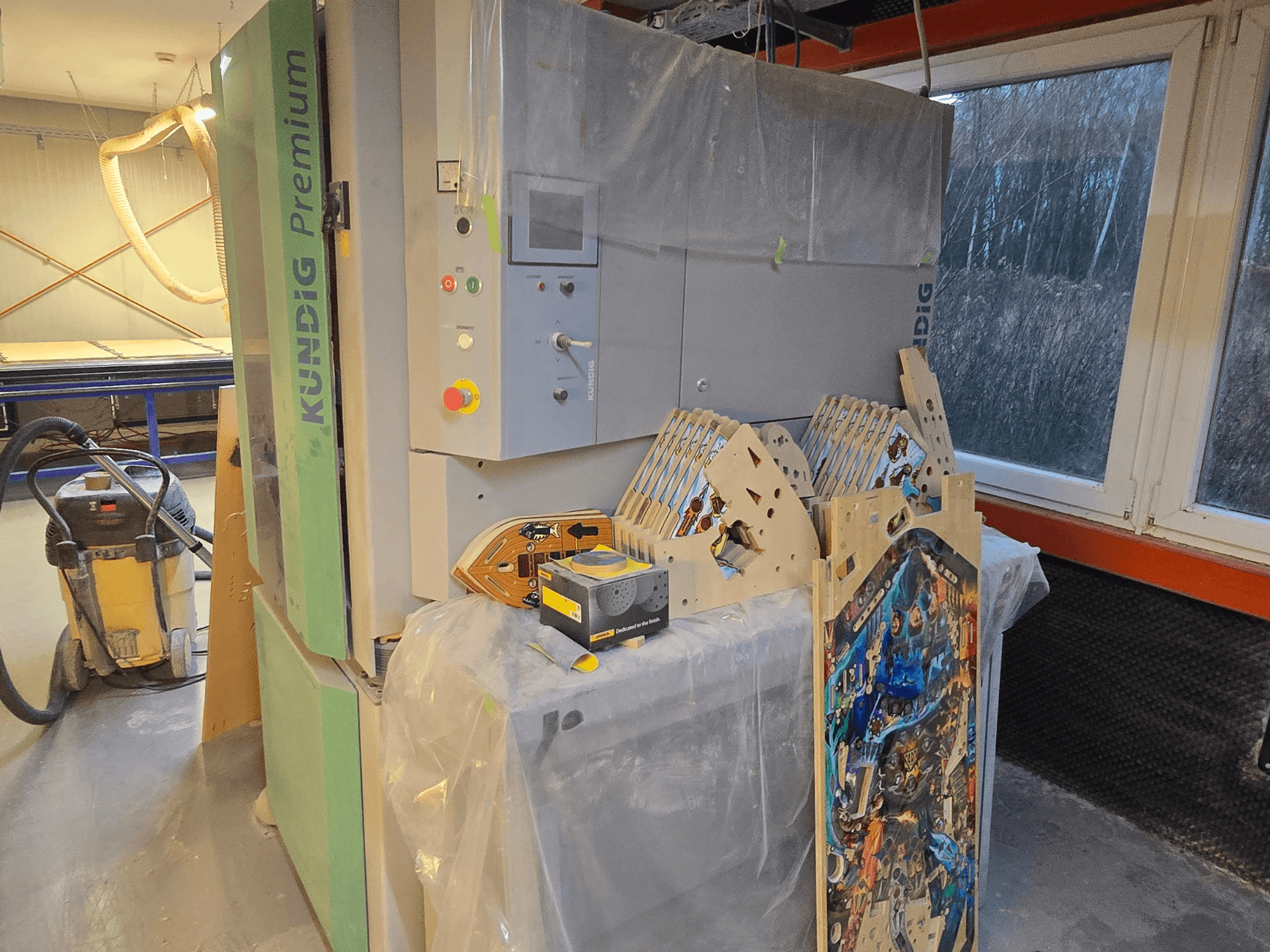 Kundig Premium-1 1100-C bandschuurmachine in de werkplaats, met bedieningspaneel en houtstukken, zijaanzicht, met stofzuiger op de achtergrond.