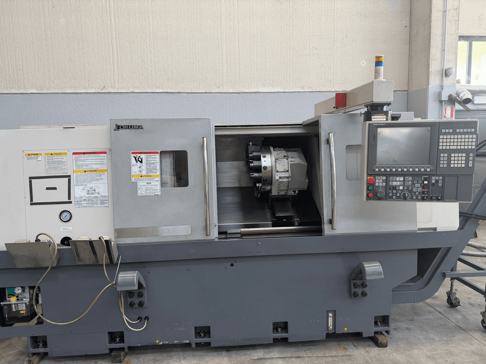 Okuma CNC draaibank, vooraanzicht, met bedieningspaneel, houder voor snijgereedschap en veiligheidswaarschuwingen op de zijkant.