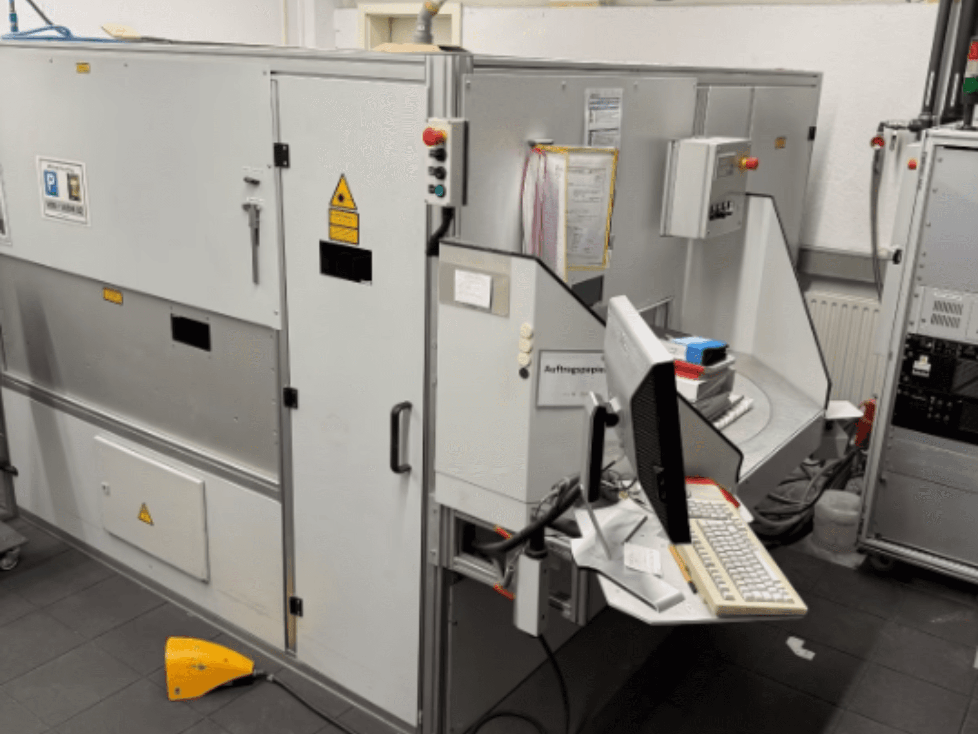 Industriële machine met een computerterminal, zijaanzicht; inclusief bedieningsknoppen en veiligheidswaarschuwingstekens.