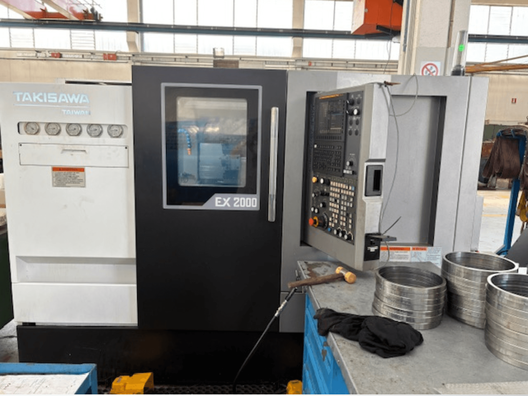 Takisawa EX 2000 CNC machine, vooraanzicht, met bedieningspaneel, meters en metalen onderdelen op een werkplaatstafel.