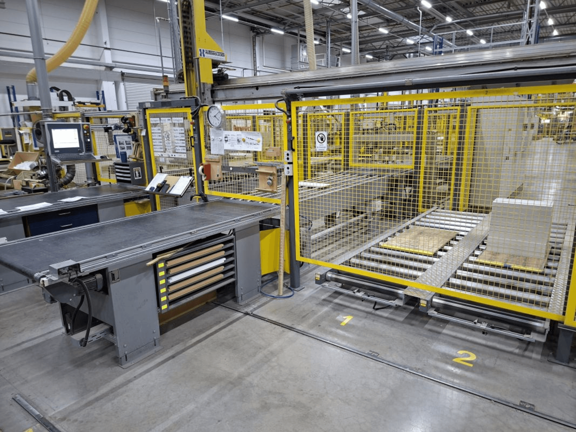 Industriële werkruimte met een transportsysteem en bedieningspaneel; zijaanzicht met machines en veiligheidshekken.