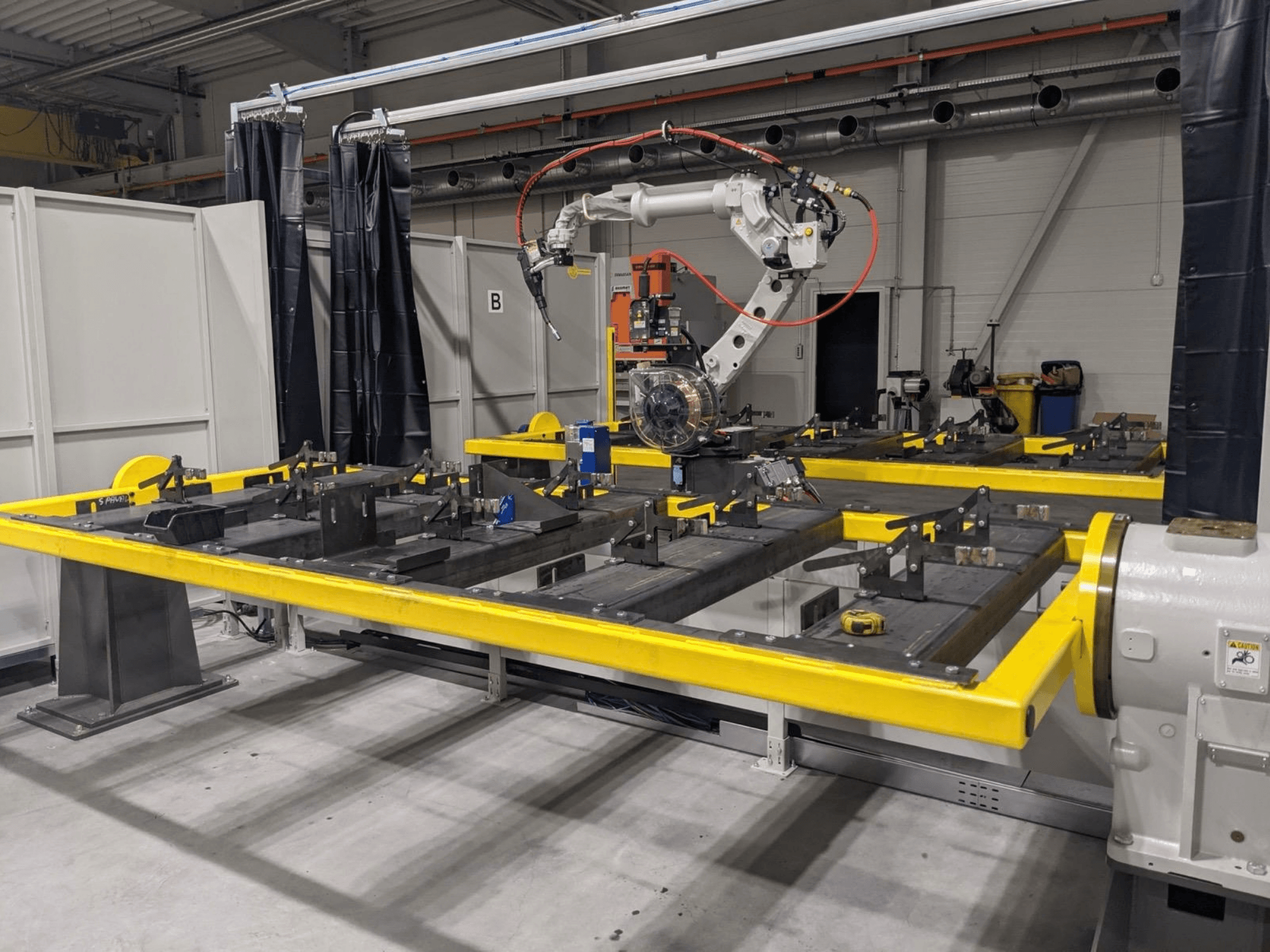 Vooraanzicht van de PANASONIC TAWERS TM-2000WG3-robotarm, geplaatst boven een gele werktafel met metalen onderdelen die gemonteerd moeten worden.